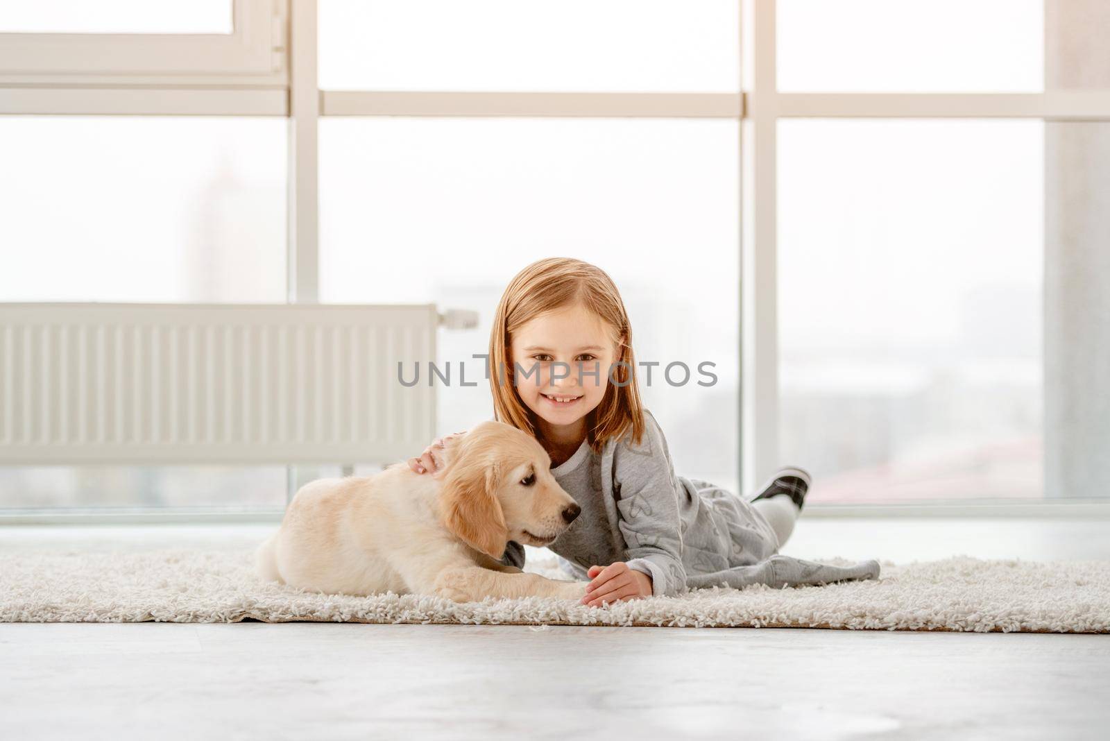 Smiling little girl with young dog golden retriever lying in light room