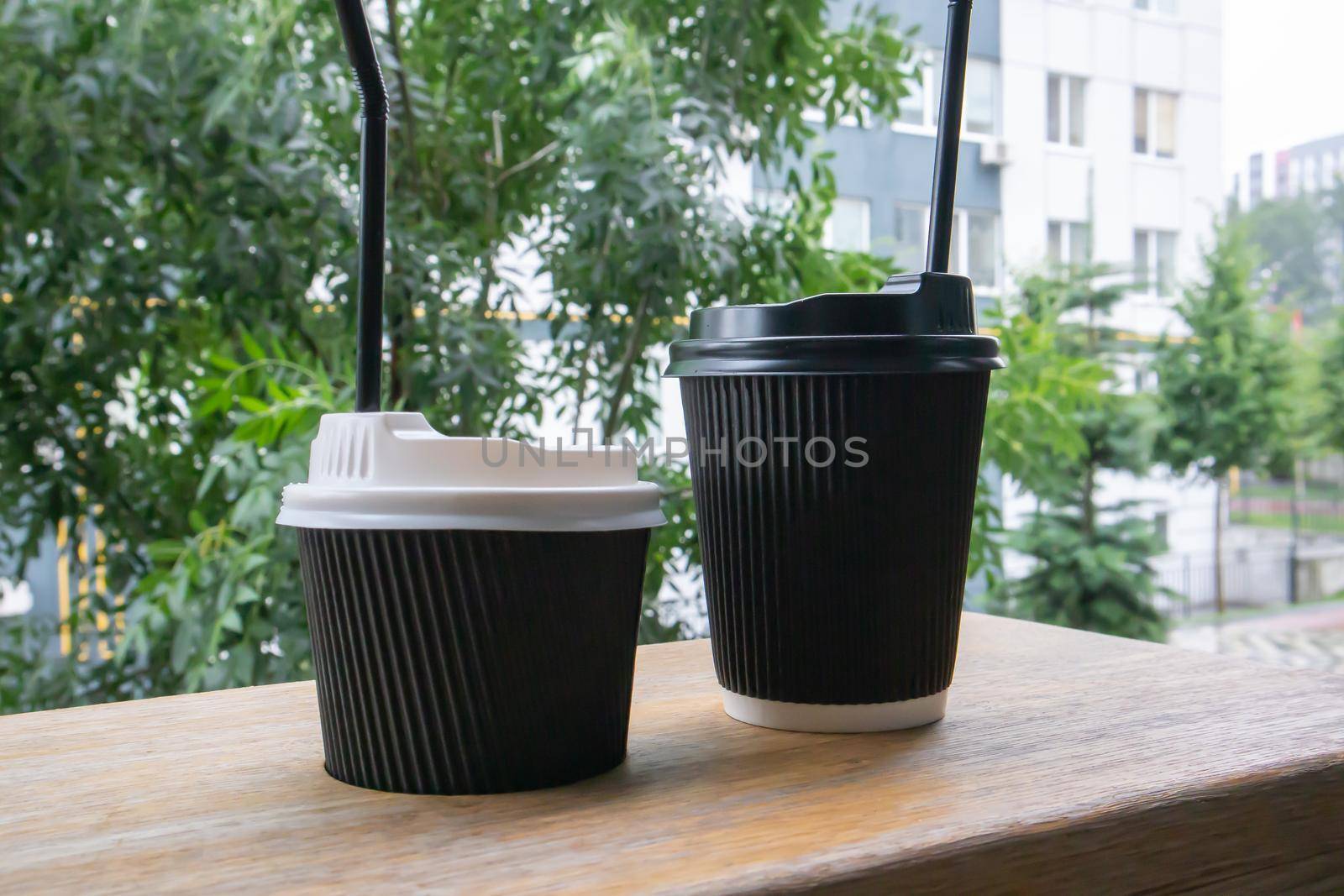 Two black paper coffee cups with lids outdoors in summer in sunny weather on a wooden table of a cafe, coffee shop or restaurant