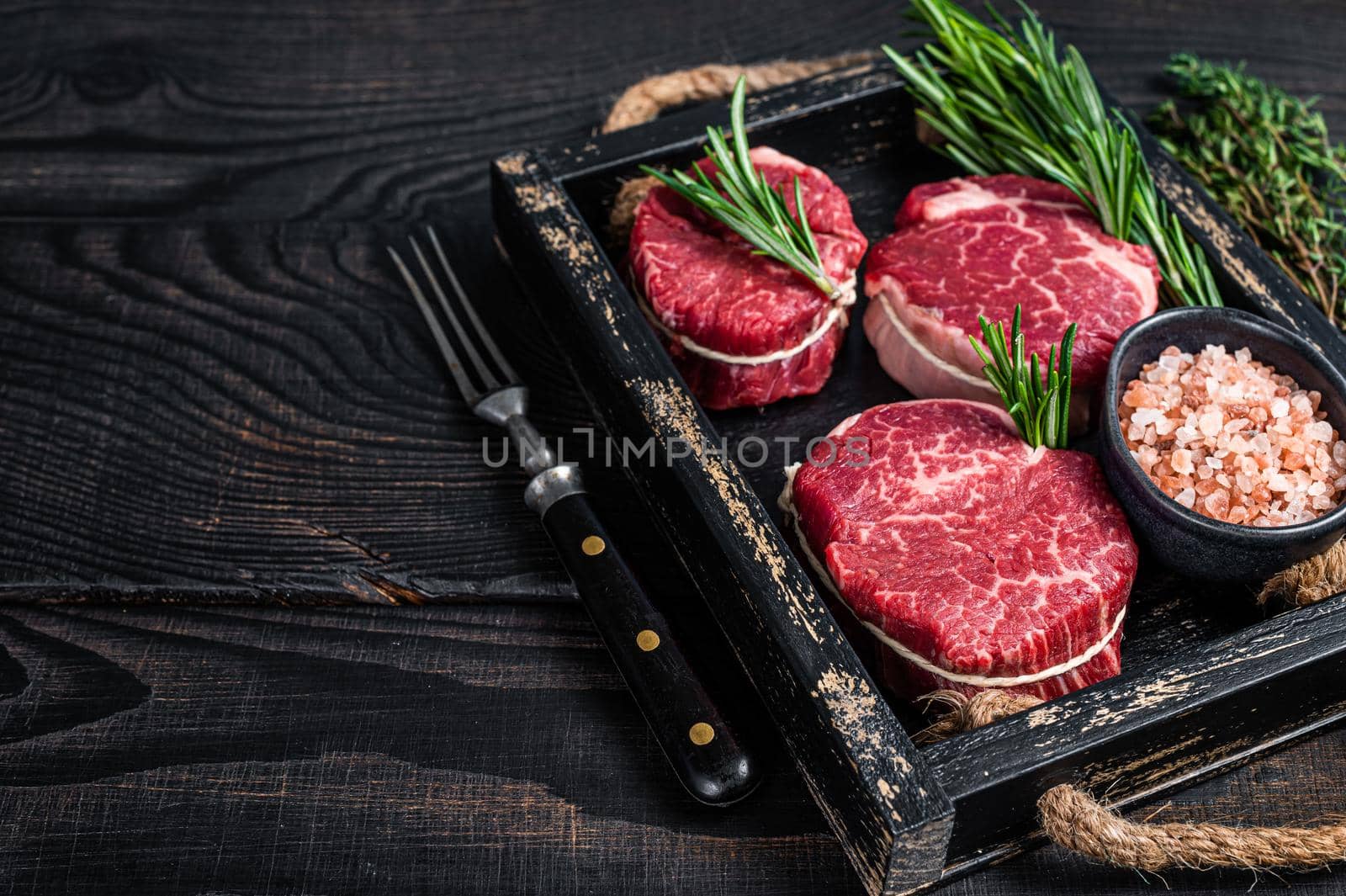 Fillet Mignon tenderloin raw meat veak steaks in wooden tray with herbs. Black wooden background. Top view. Copy space by Composter