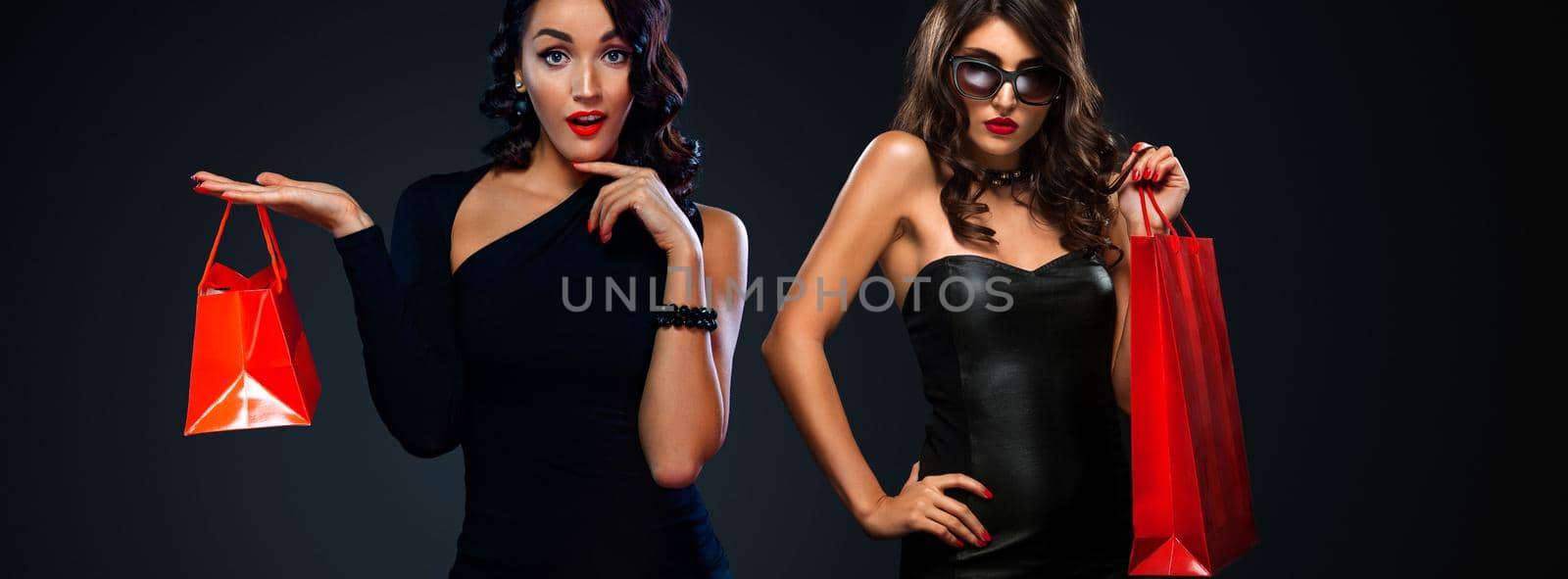 Black Friday sale concept for shops. Two woman in sunglasses holding red bag isolated on dark background at shopping mall. by MikeOrlov