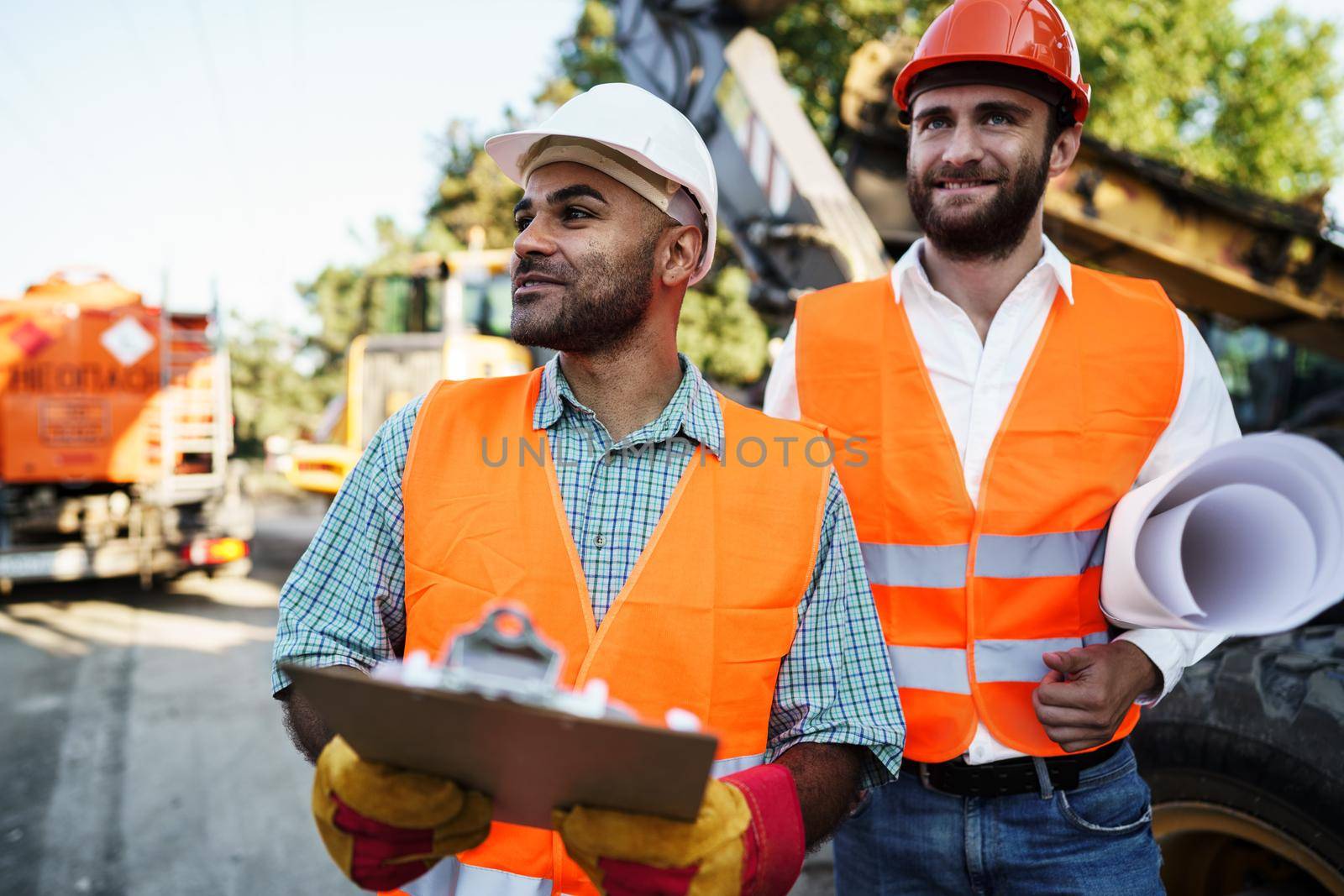 Two men engineers in workwear discussing their work standing against construction machines