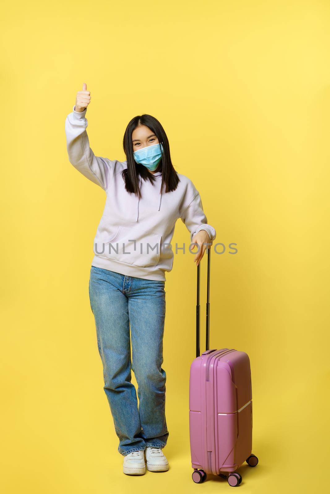 Full length shot of enthusiastic korean girl enjoying vacation, posing with suitcase, wearing face medical mask, travelling abroad during covid pandemic, yellow background.