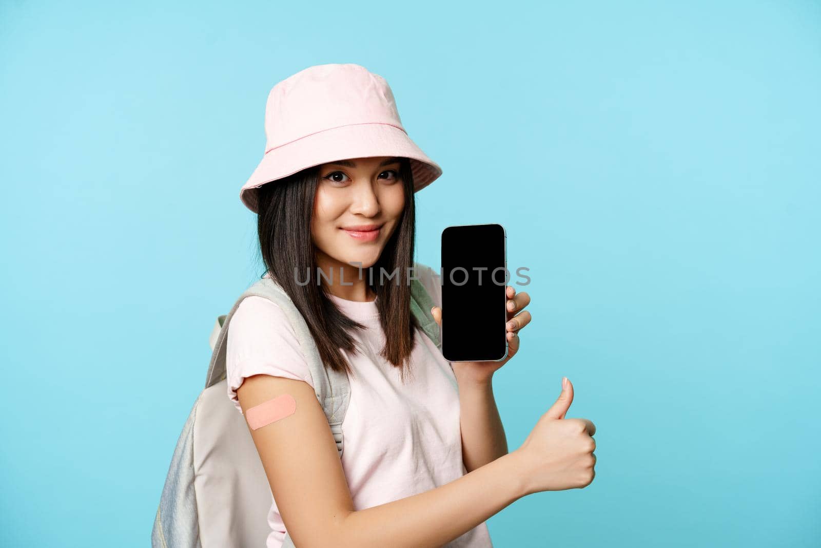 Happy asian woman showing health passport on mobile phone screen, vaccinated hand with patch, thumbs up. Tourist made covid-19 vaccine for international certificate, blue background.