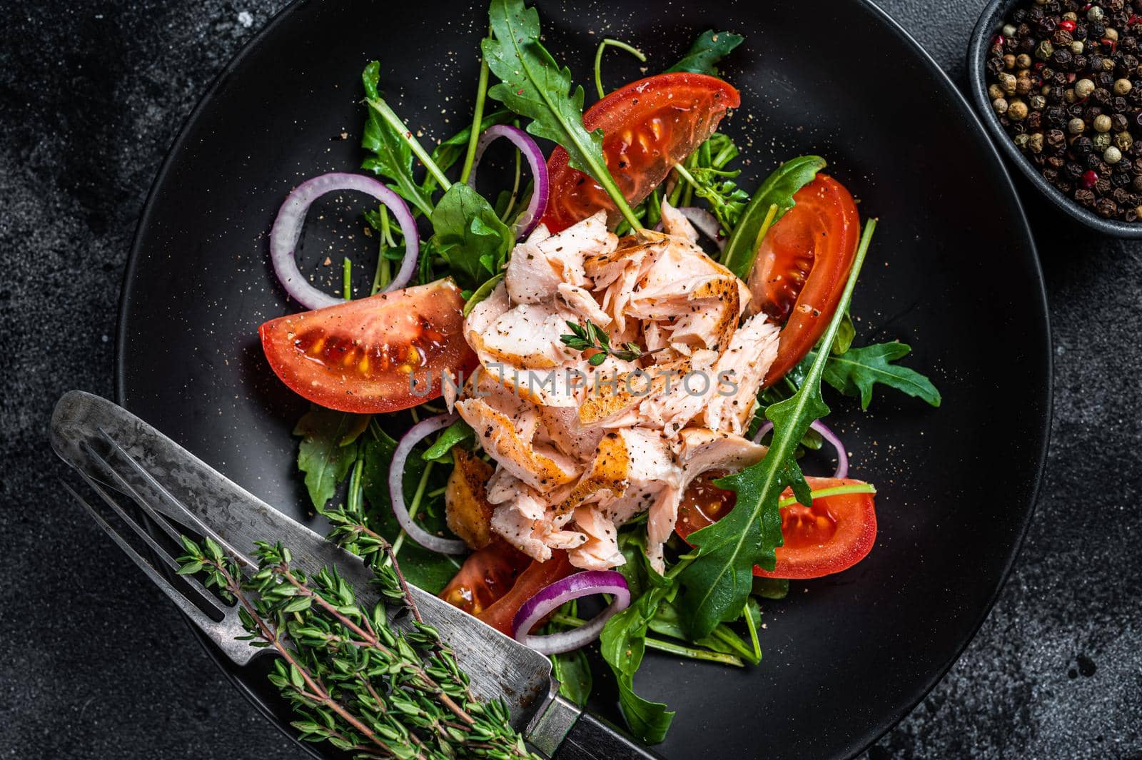 Salad with baked salmon fillet steak, fresh arugula and tomato in a plate. Black background. Top view.