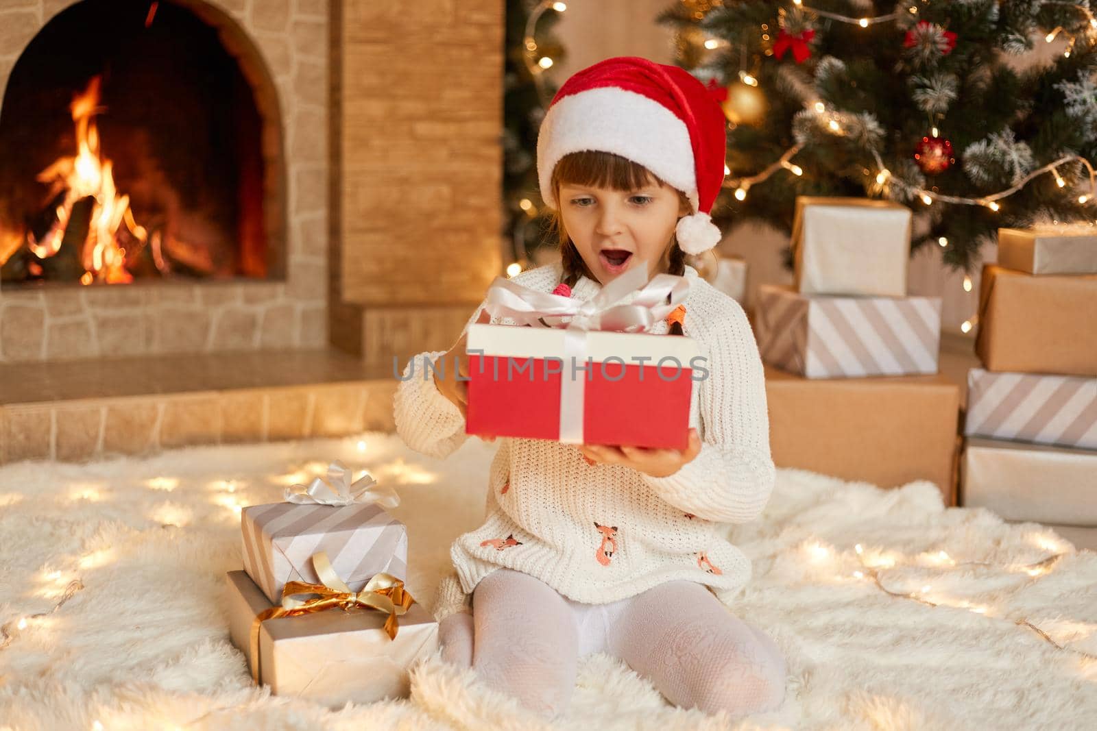 Surprised little girl wearing santa Claus hat and white warm sweater sitting on floor with gift box, look at her present with big eyes and opened mouth, being excited and astonished. by sementsovalesia