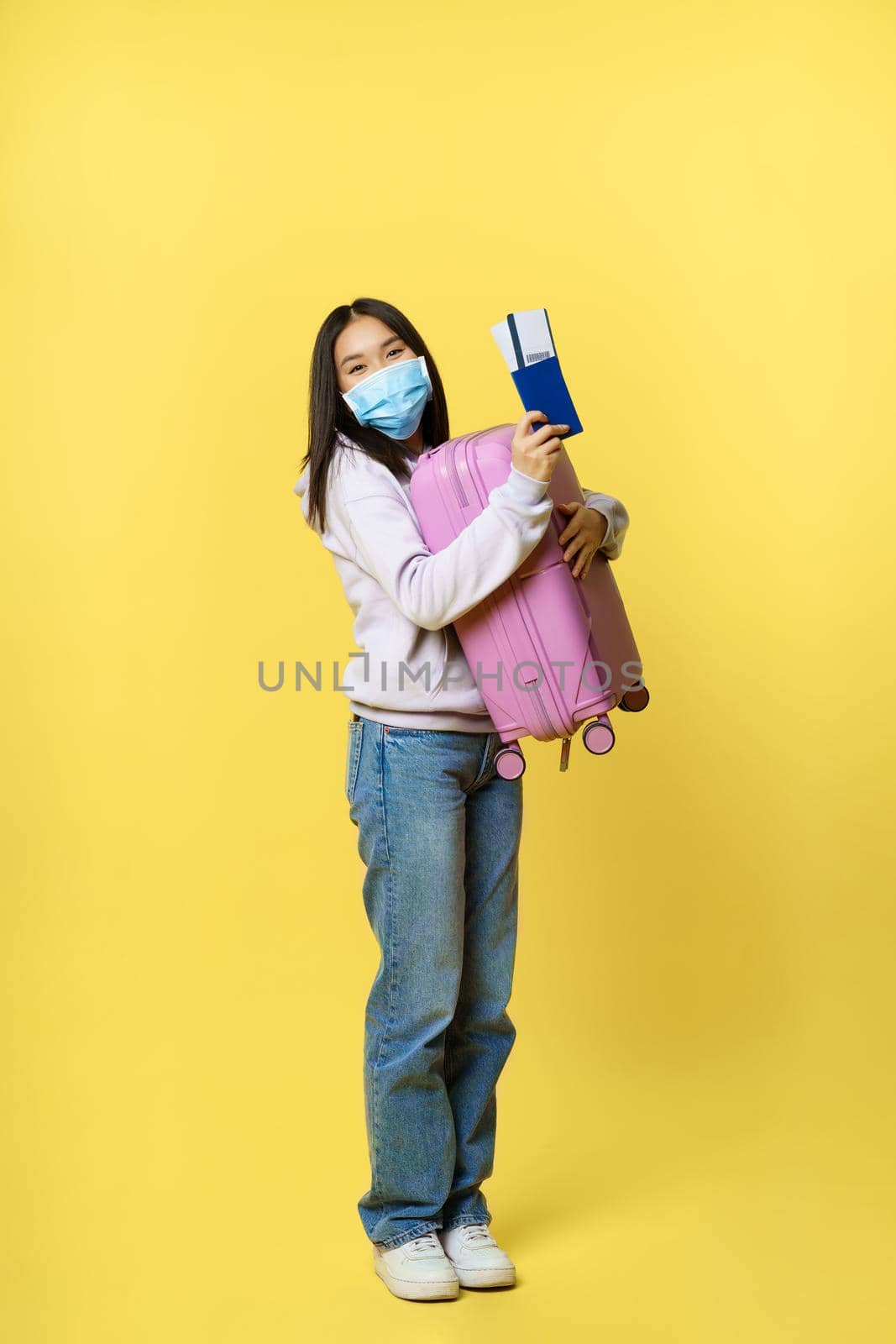Full length shot cute korean girl tourist in medical face mask, holding her suitcase and passport with flight tickets, going on vacation abroad, yellow background.