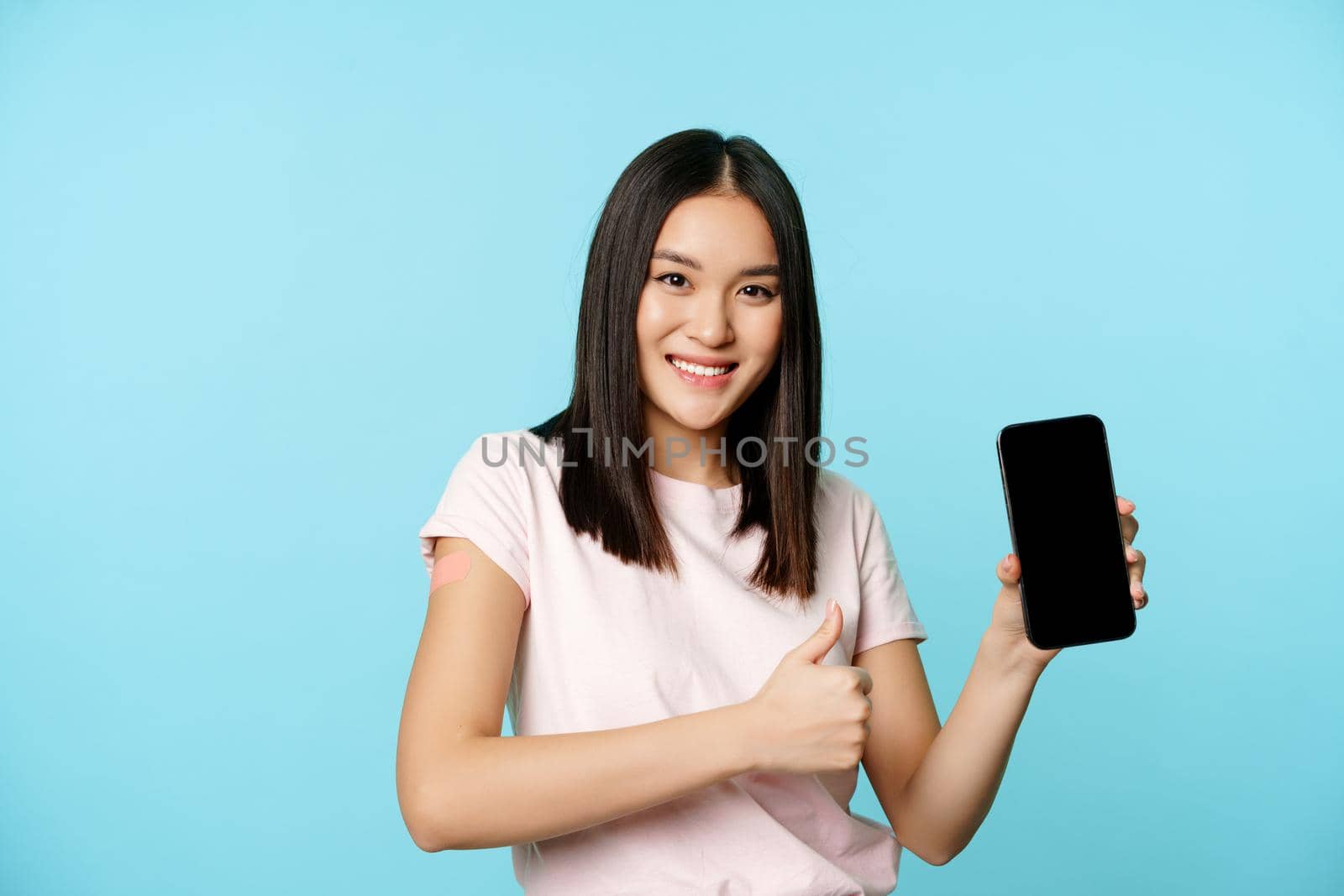 Smiling asian girl got vaccinate from coronavirus, showing thumbs up and smartphone app interface, concept of health passport, international vaccination certificate from covid.