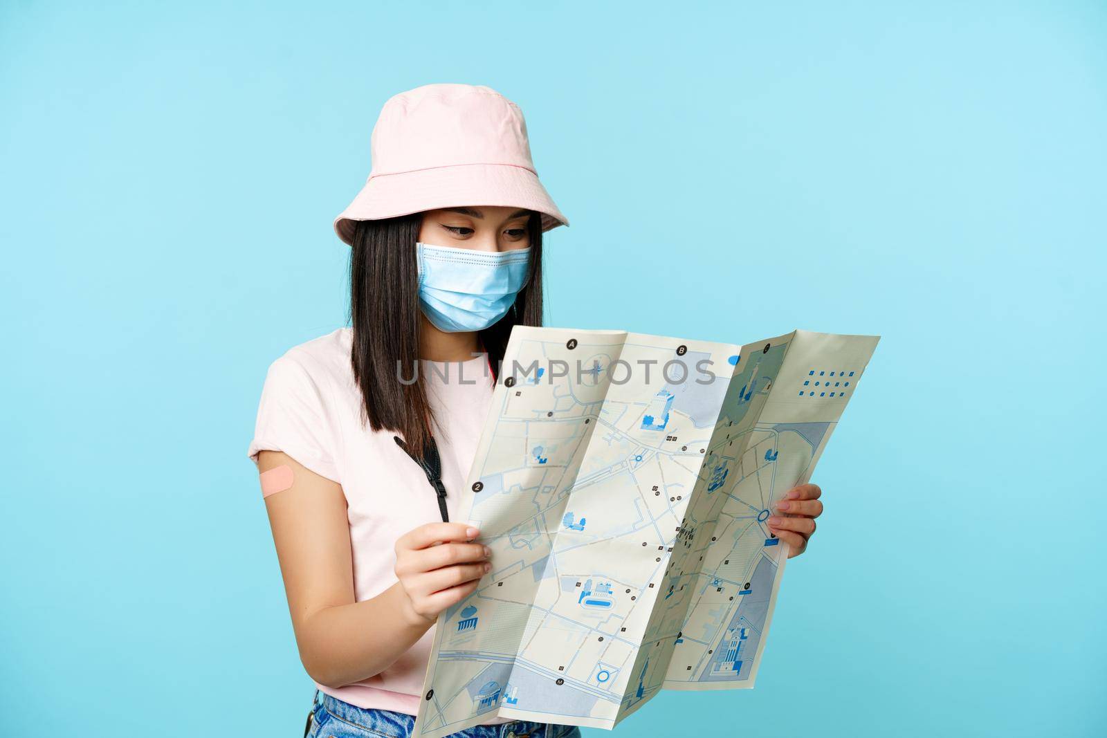 Asian girl tourist in medical face mask, vaccinated from covid with patch on arm, travelling abroad, looking at sightseeing map, exploring tourism spots, standing over blue background.