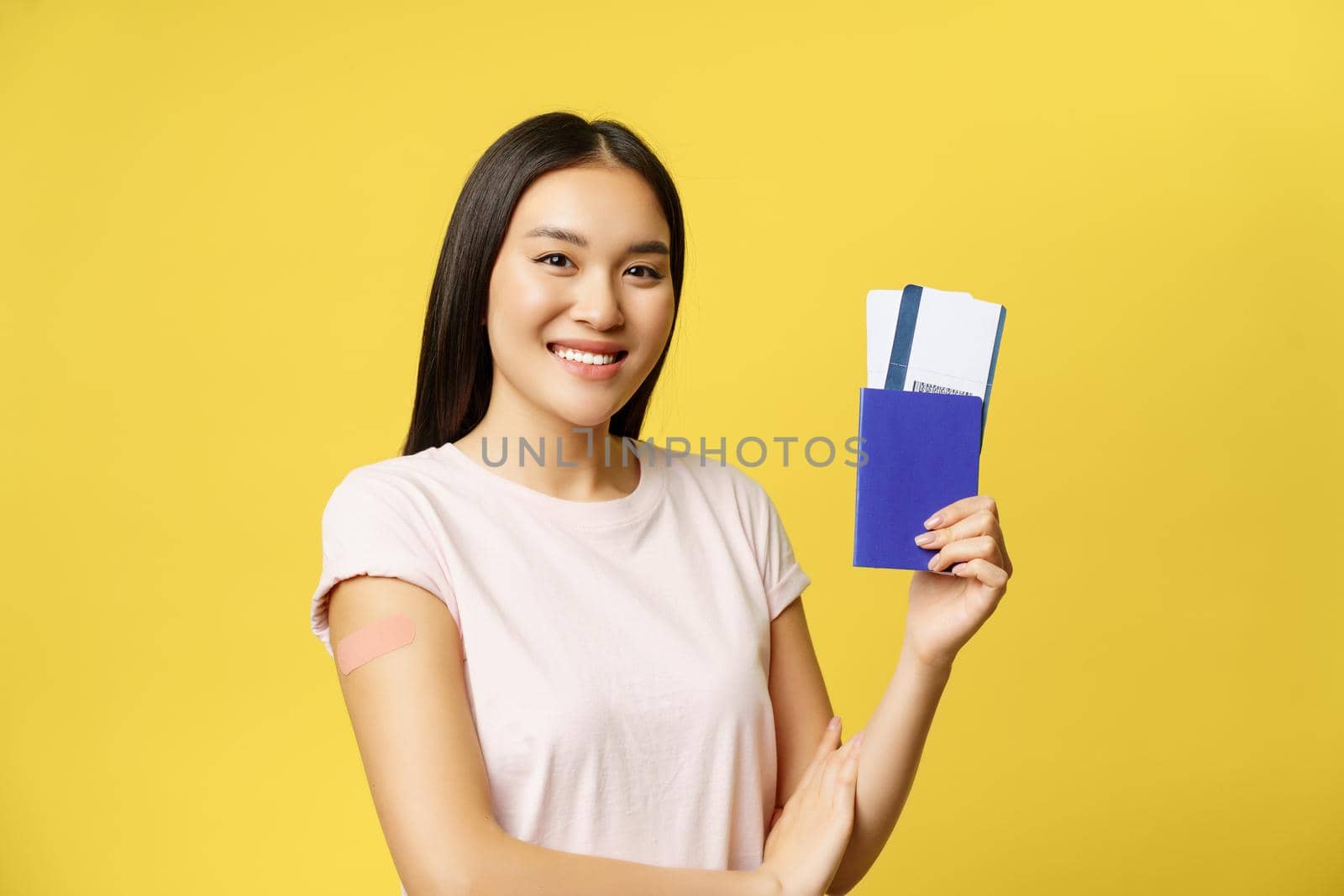 Smiling asian woman showing passport with tickets, arm with patch after covid 19 vaccination, vaccinating campaign for tourists, yellow background.