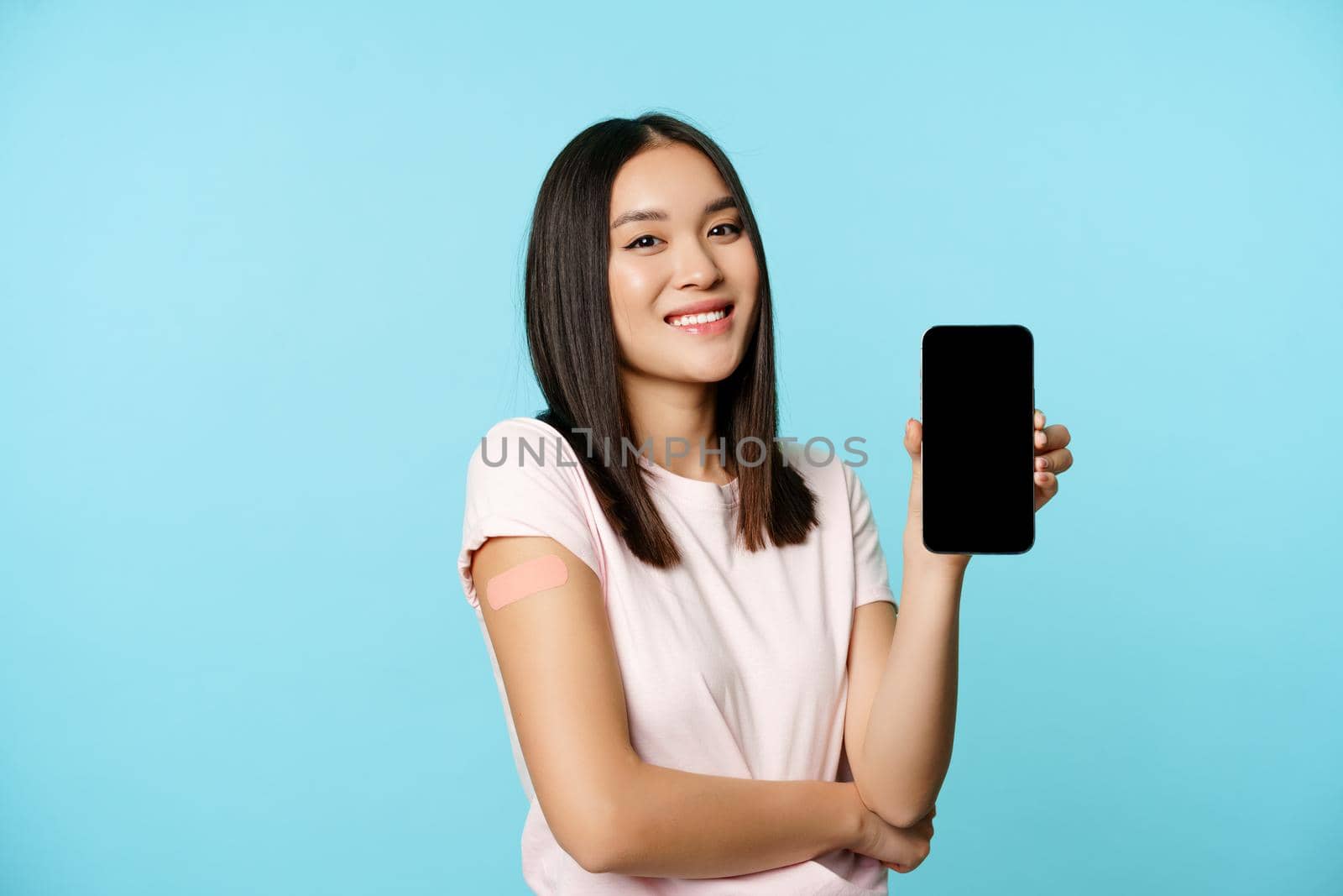 Smiling asian girl with vaccinated arm, showing mobile phone empty screen, concept of health passport, covid-19 vaccination certificate on smartphone.