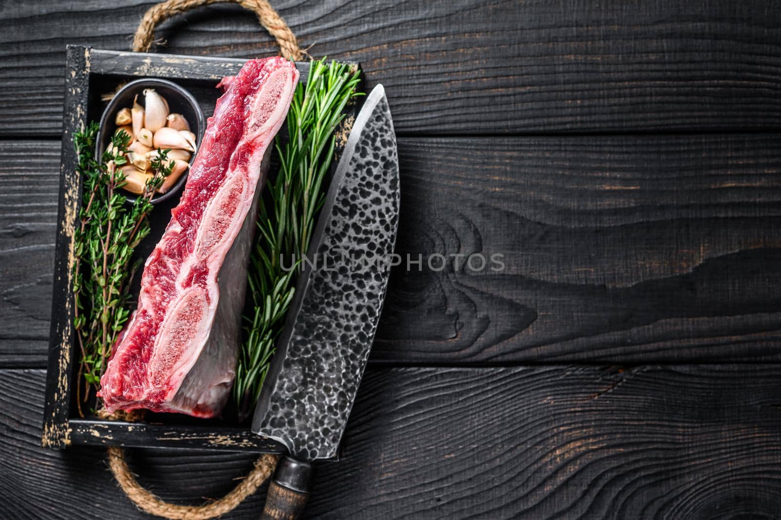 Fresh Raw veal short ribs in a wooden tray with herbs. Black wooden background. Top view. Copy space.