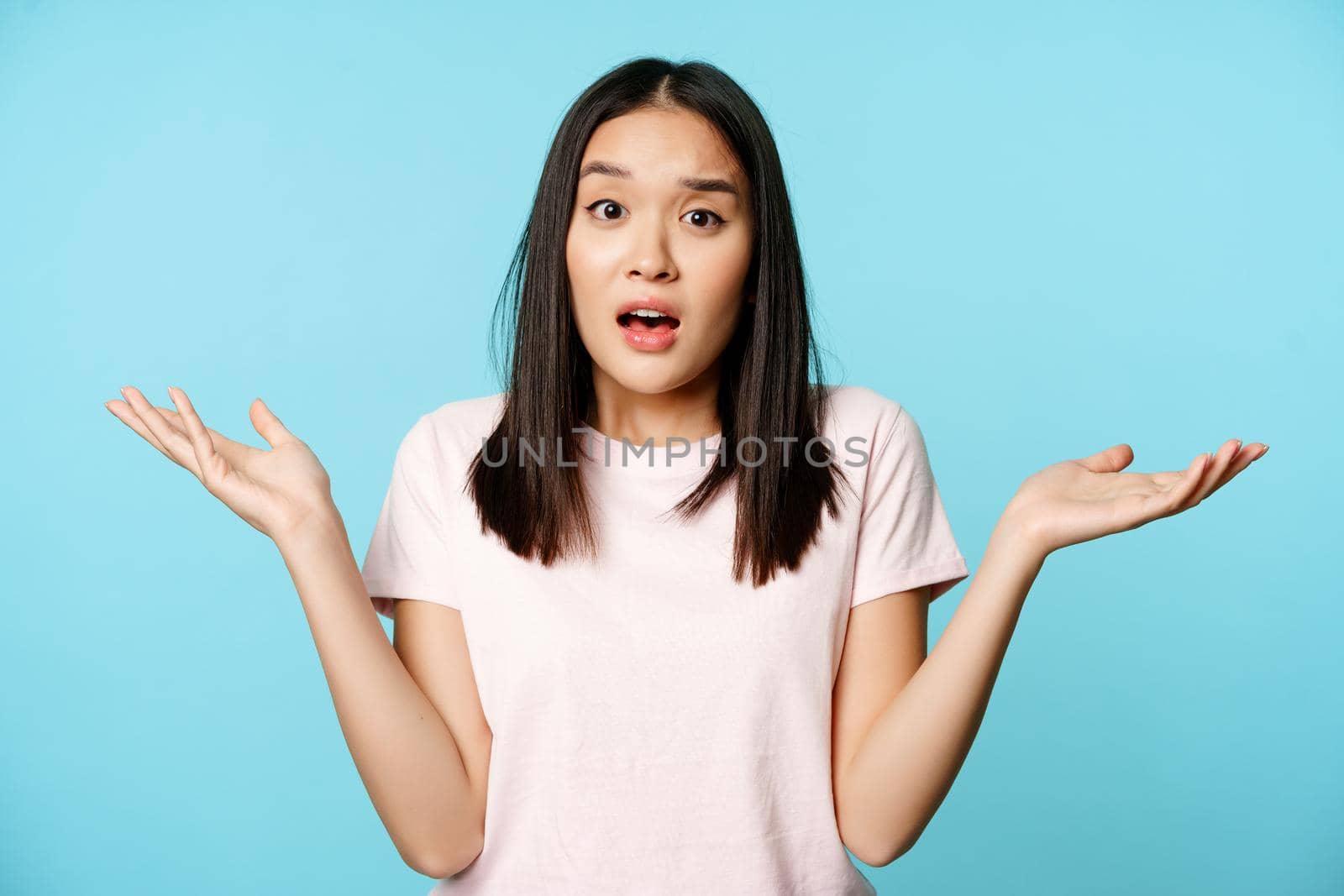 Confused asian girl spread empty hands sideways, shrugging shoulders clueless, standing in tshirt over blue background by Benzoix