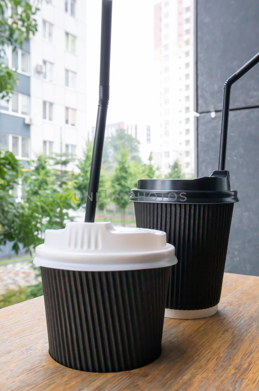Two black paper coffee cups with lids outdoors in summer in sunny weather on a wooden table of a cafe, coffee shop or restaurant