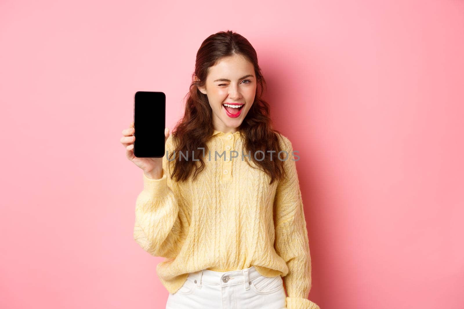 Image of cheeky attractive girl, showing you smartphone screen, winking and smiling, recommending mobile phone, standing against pink background by Benzoix