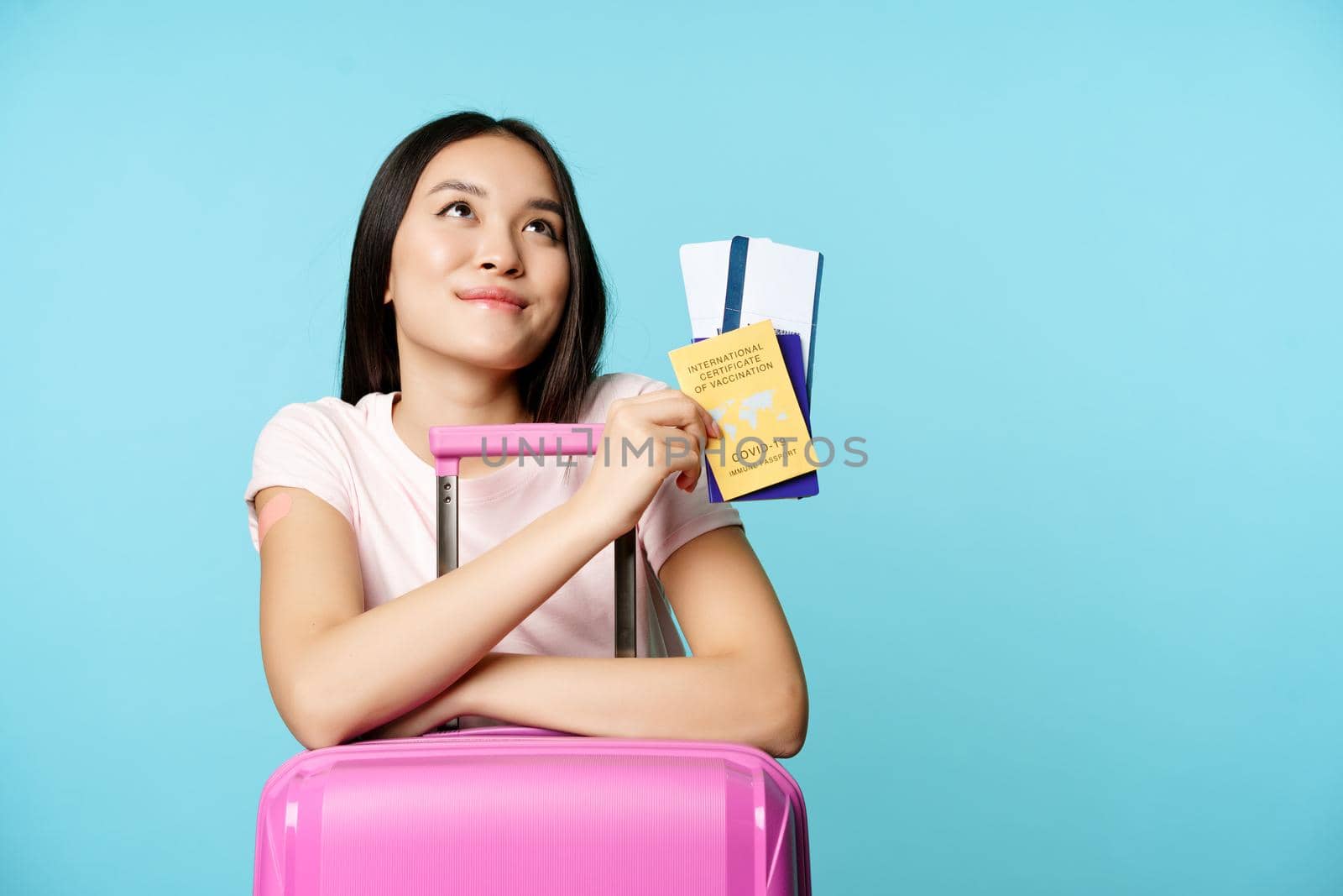 Happy asian female tourist dreaming of trip, lean on suitcase, holding covid-19 international vaccination certificate, two tickets and passport, thinking of journey, blue background.
