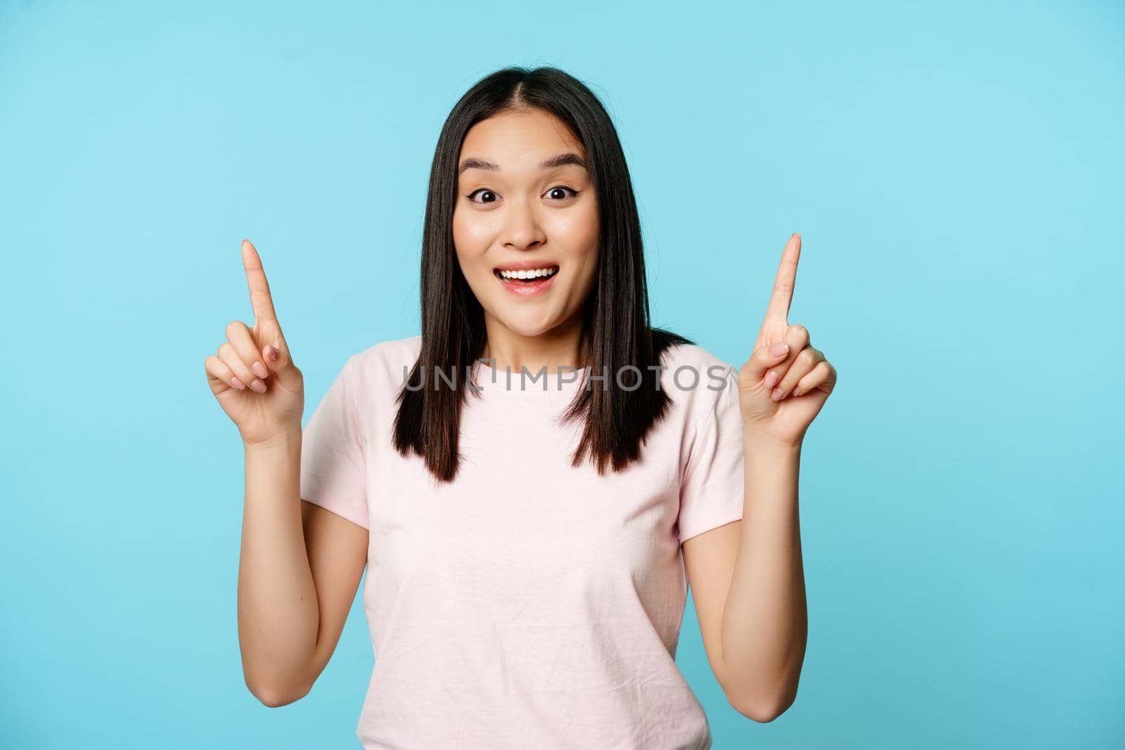 Enthusiastic korean girl shows advertisement, points up and smiles amazed, checks out promo deal, stands over blue background.