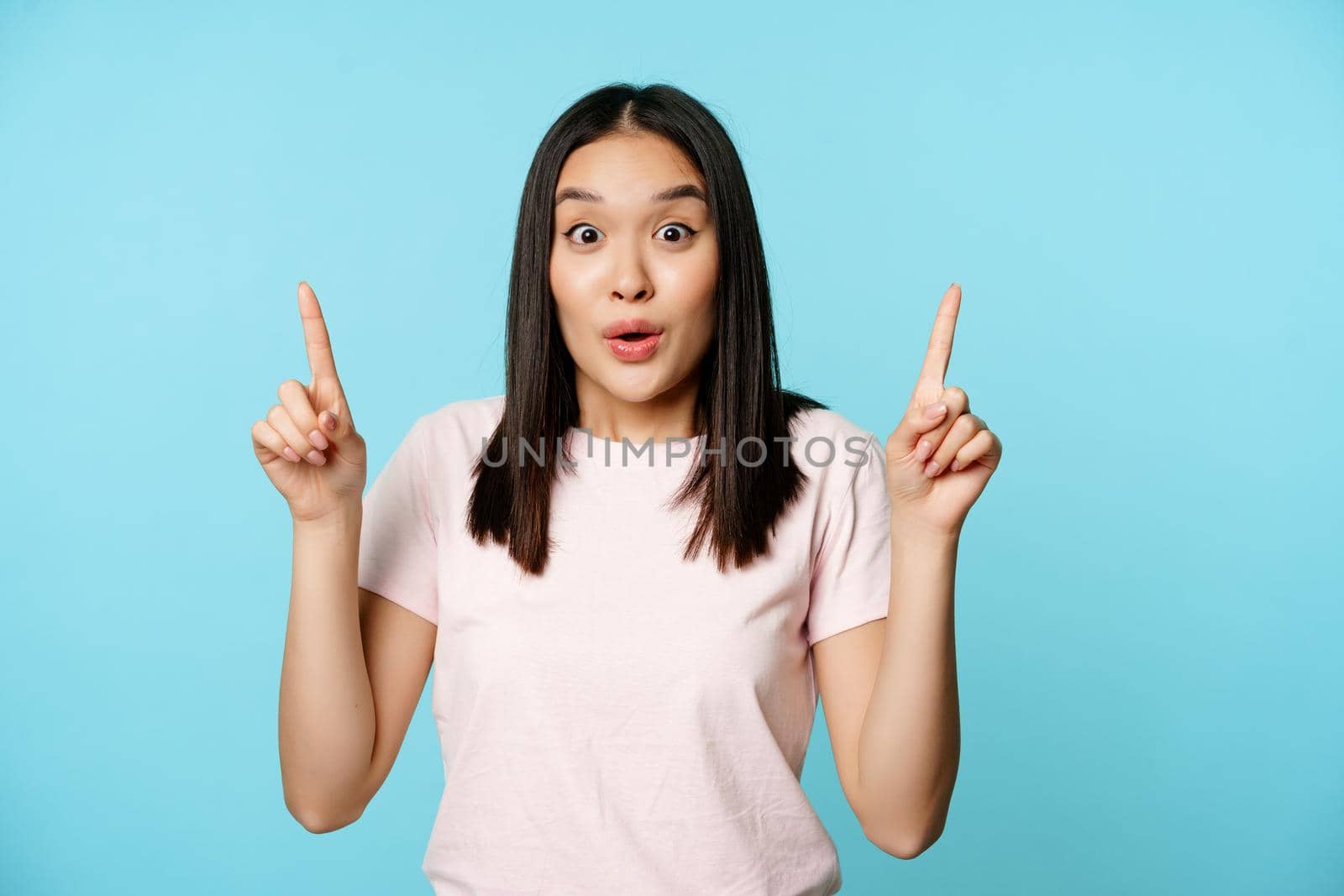 Portrait of surprised korean woman gasp in awe, pointing fingers up, showing advertisement, standing over blue background.
