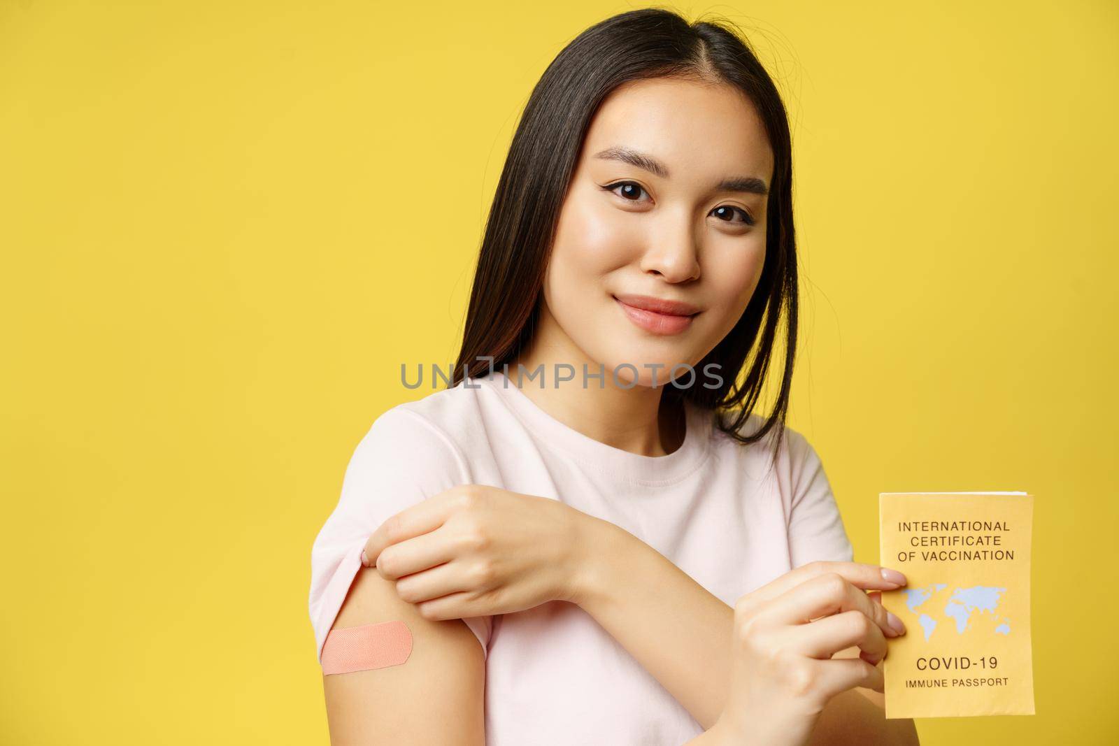 Covid-19 and tourism concept. Portrait of young asian woman showing her patch from covid 19 vaccine, holding coronavirus international vaccination passport, yellow background.