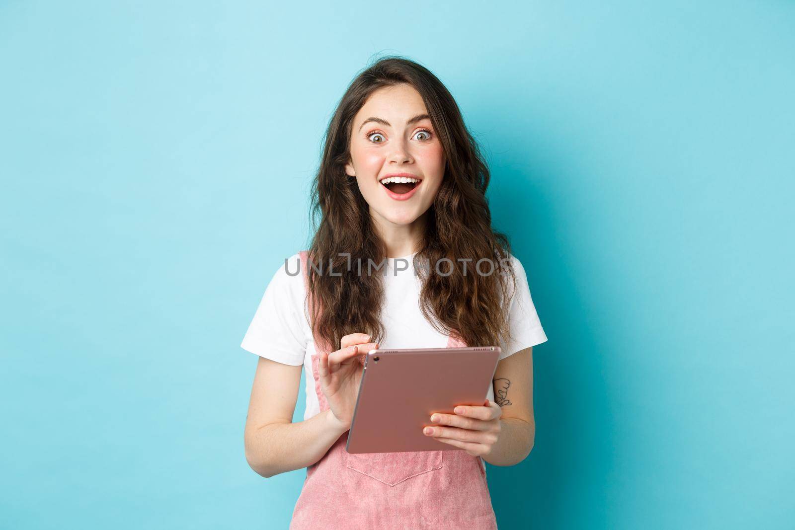 Excited smiling woman holding digital tablet, staring amazed at camera after seeing cool offer online, standing against blue background.