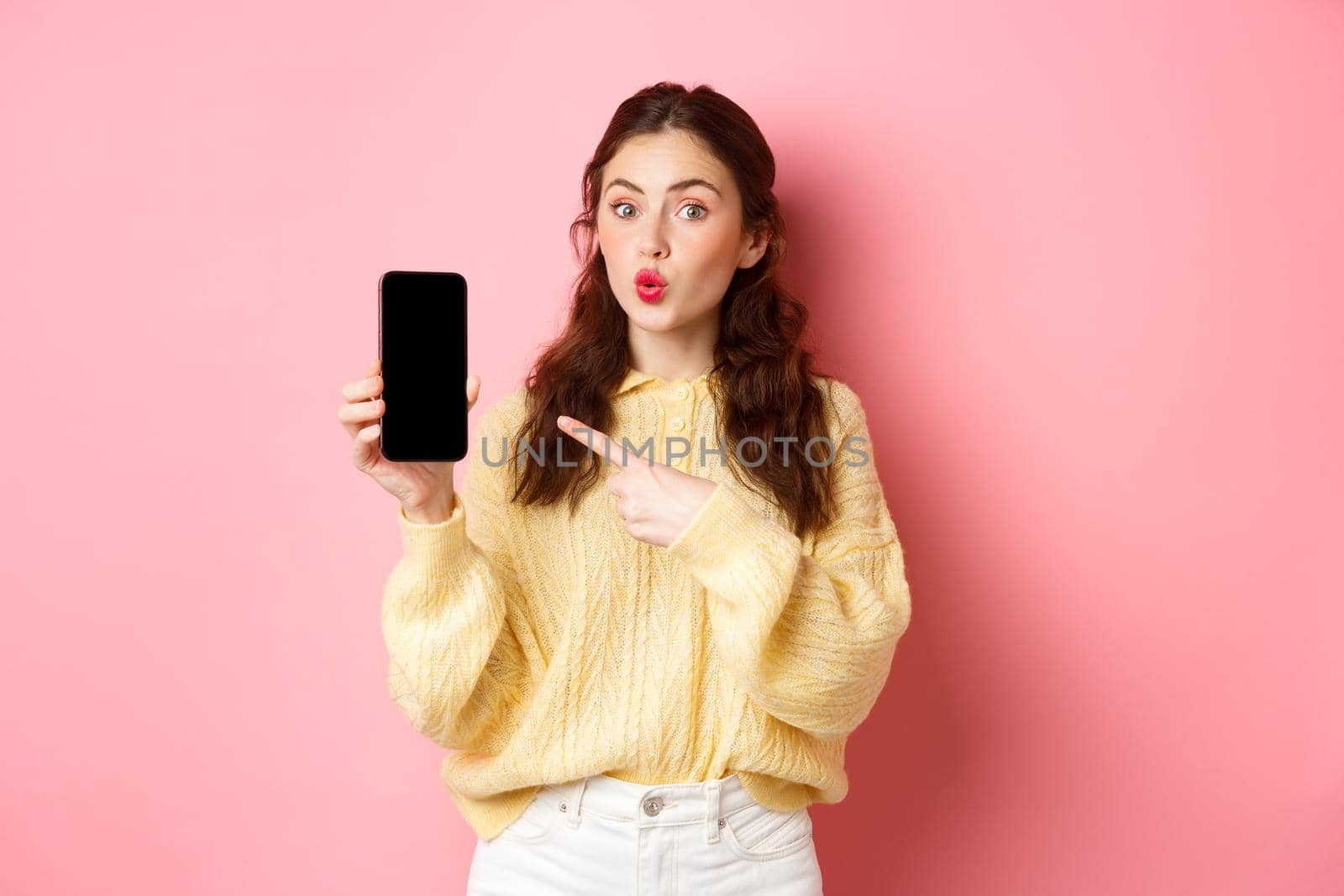 Technology and online shopping. Excited girl looks curious, points finger at empty mobile phone screen, shows app on smartphone, standing against pink background.