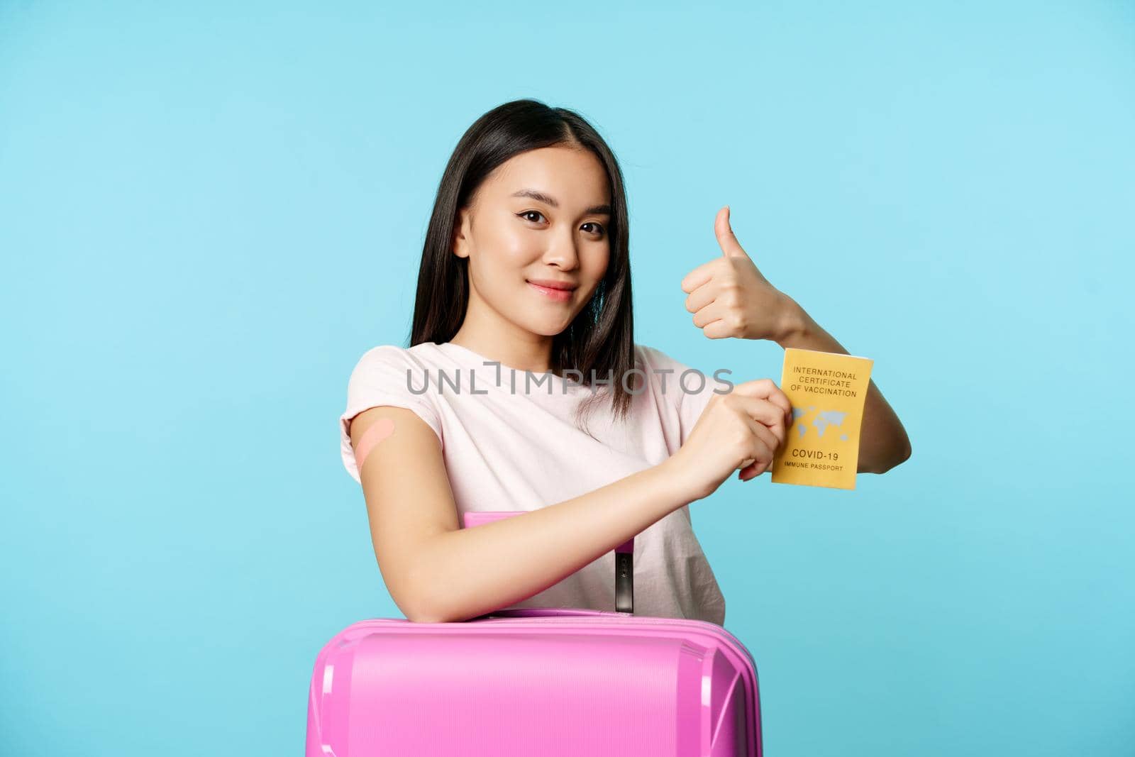 Healthcare and tourism. Smiling asian girl got vaccinated from covid-19, standing with suitcase, showing coronavirus international vaccination passport, thumbs up and arm with patch by Benzoix