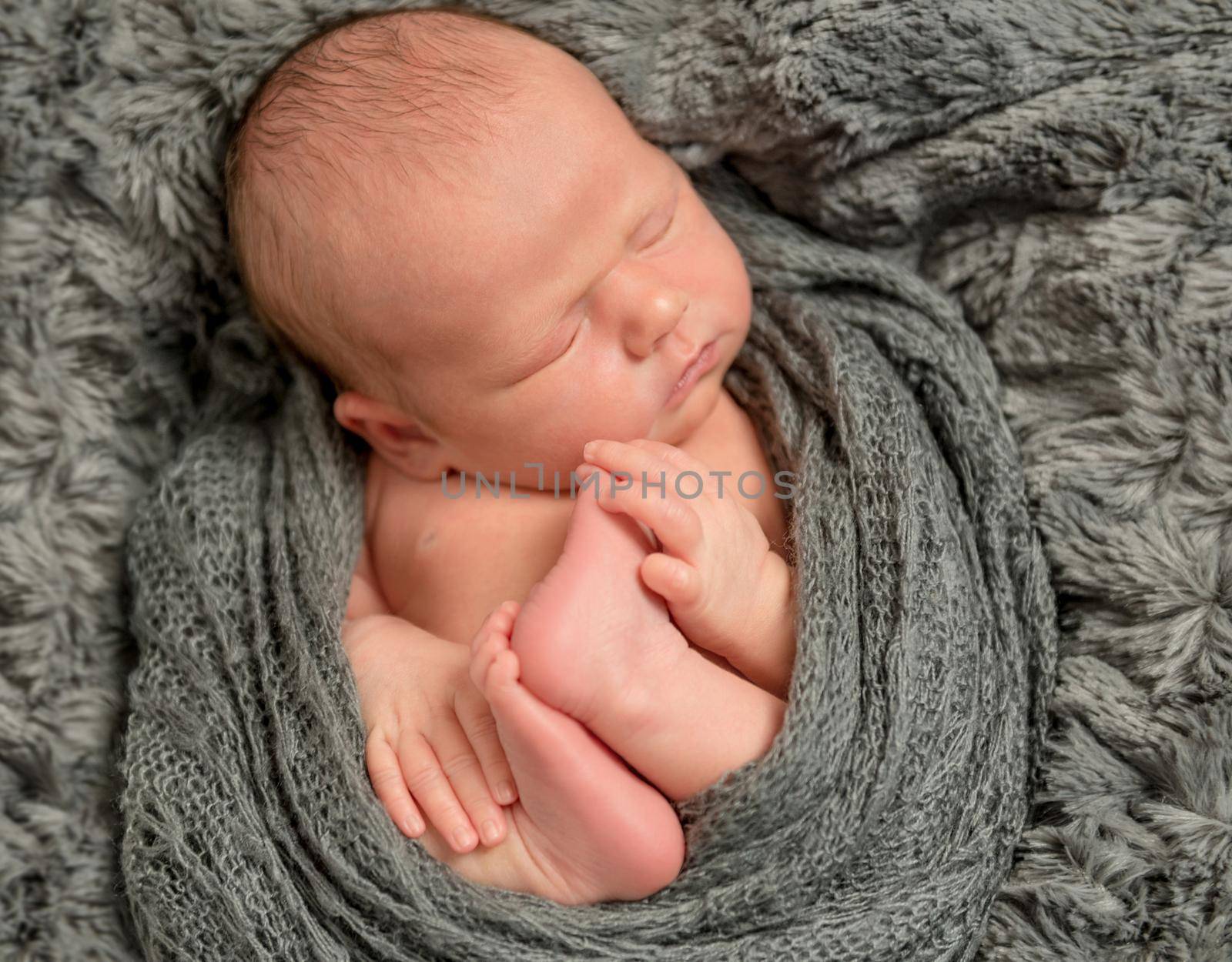 Adorable infant holding his feet in a sleep, enveloped with grey blanket, closeup