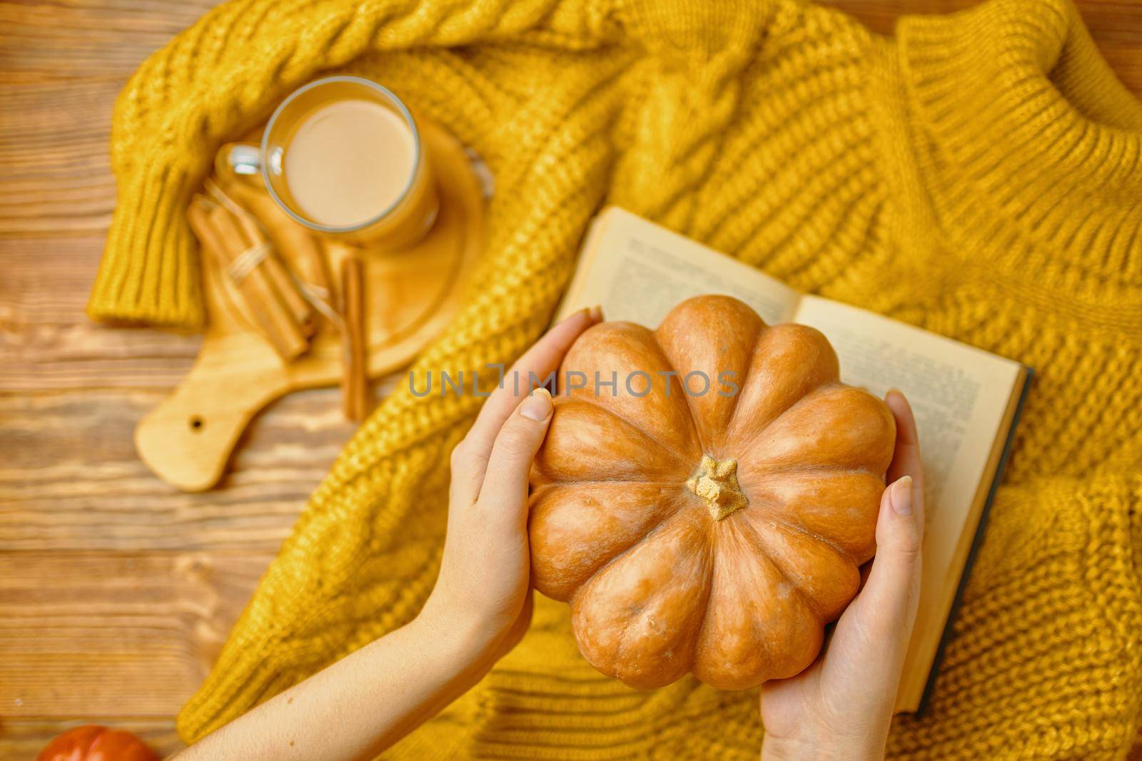 Aromatic coffee with cinnamon sticks on wooden tray. Orange sweater, open book and pumpkin on wooden background. Romantic composition, top view. Round pumpkin in hands over warm knitted sweater.