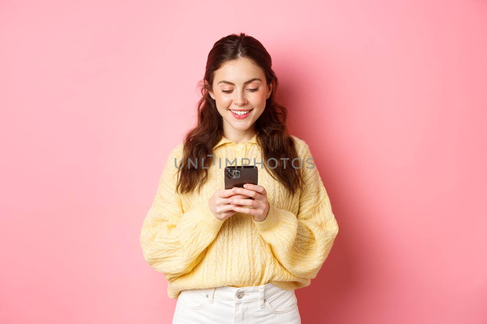 Portrait of young brunette woman reading smartphone screen and smiling, chatting on social media app, shopping online, standing against pink background by Benzoix