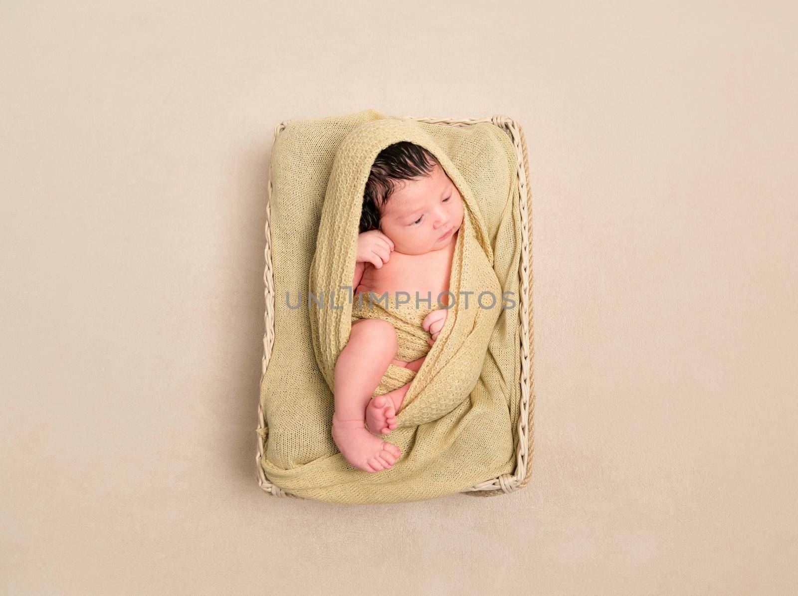 Wrapped from head to toe black-haired baby in a child's basket, resting with eyes opened