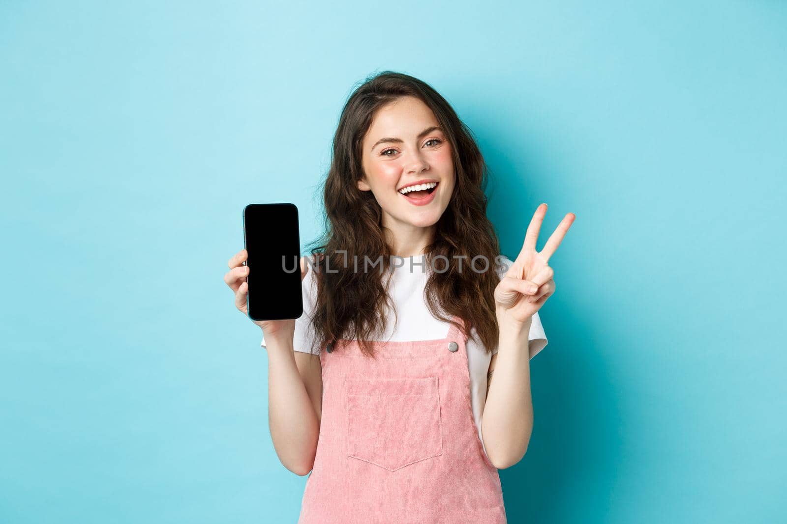 Cute young woman smiling and showing v-sign with empty smartphone screen, demonstrate app or mobile store, standing against blue background by Benzoix