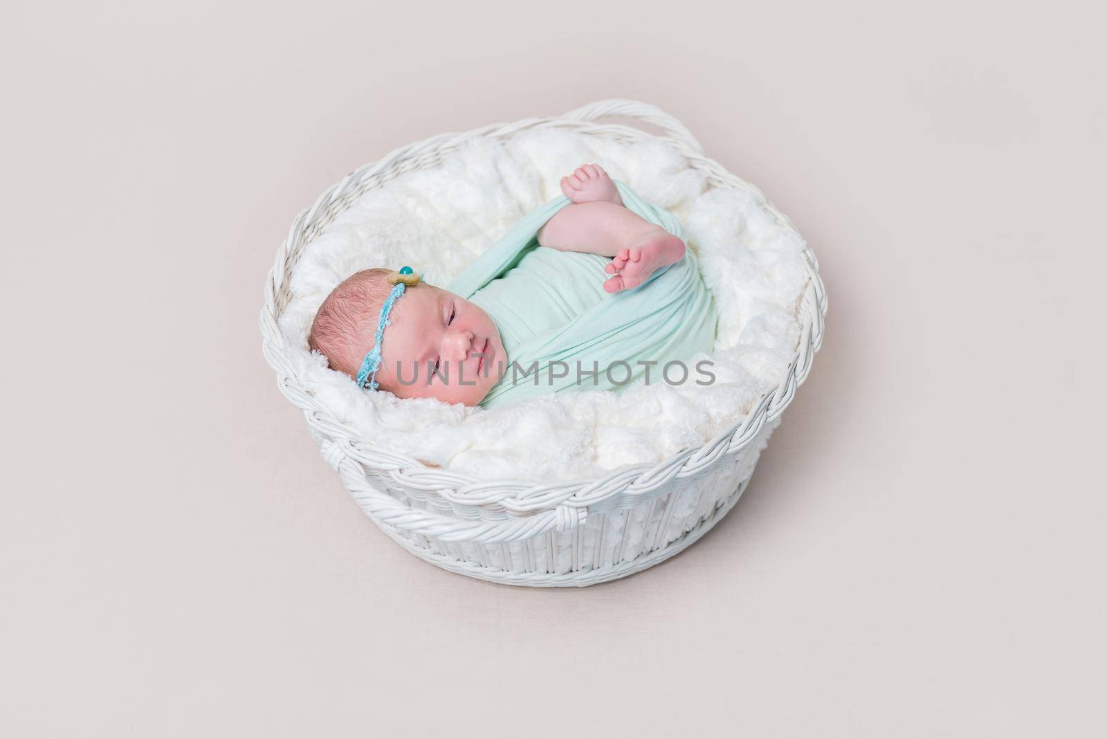 Adorable swaddled baby with legs moving, lying in kids basket, wearing headband