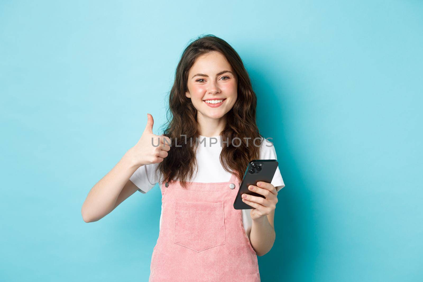 Portrait of beautiful happy girl showing thumb up and holding smartphone, online shopping, making order in internet, looking satisfied, praising good app, blue background.