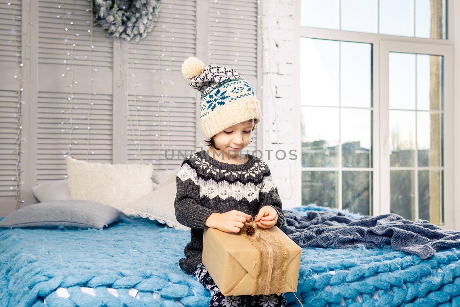 little girl the child sitting in pajamas and hat on the bed with garland of light bulbs with gifts boxes wrapped in a non-colored paper decorated with cones on blue knitted coverlet.Christmas concept.