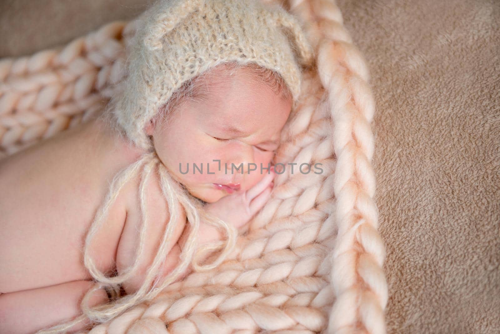 Sweetie newborn baby in beige bonnet sleeps on his tummy on knitted rug.