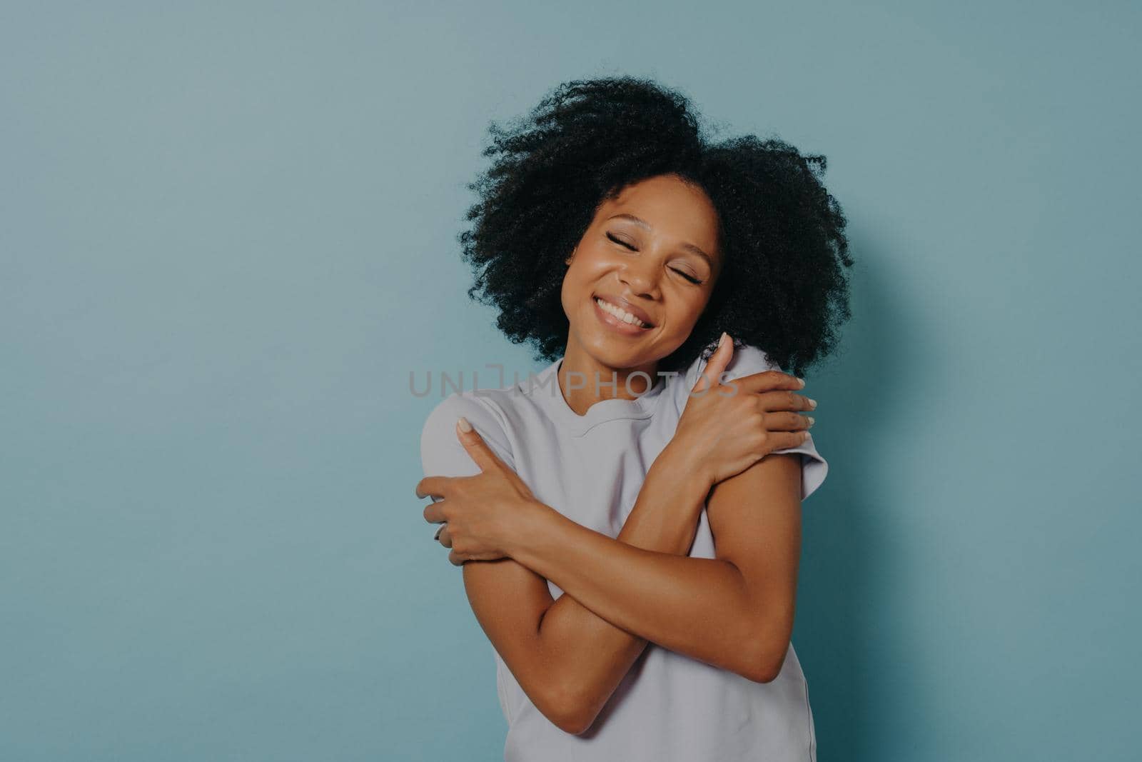 Love yourself. Happy lovely young african woman with closed eyes embracing herself, touching shoulders and tilting head while standing indoors against blue wall with copy space for text