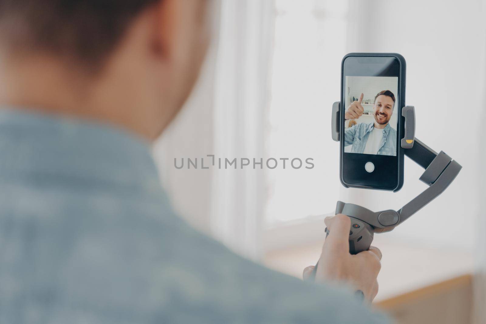 Rear view of handsome smiling young brunete man with stubble making selfie on smartphone with gimbal stabilizer at home and gesturing ok sign, wears casual clothes. Blogging concept