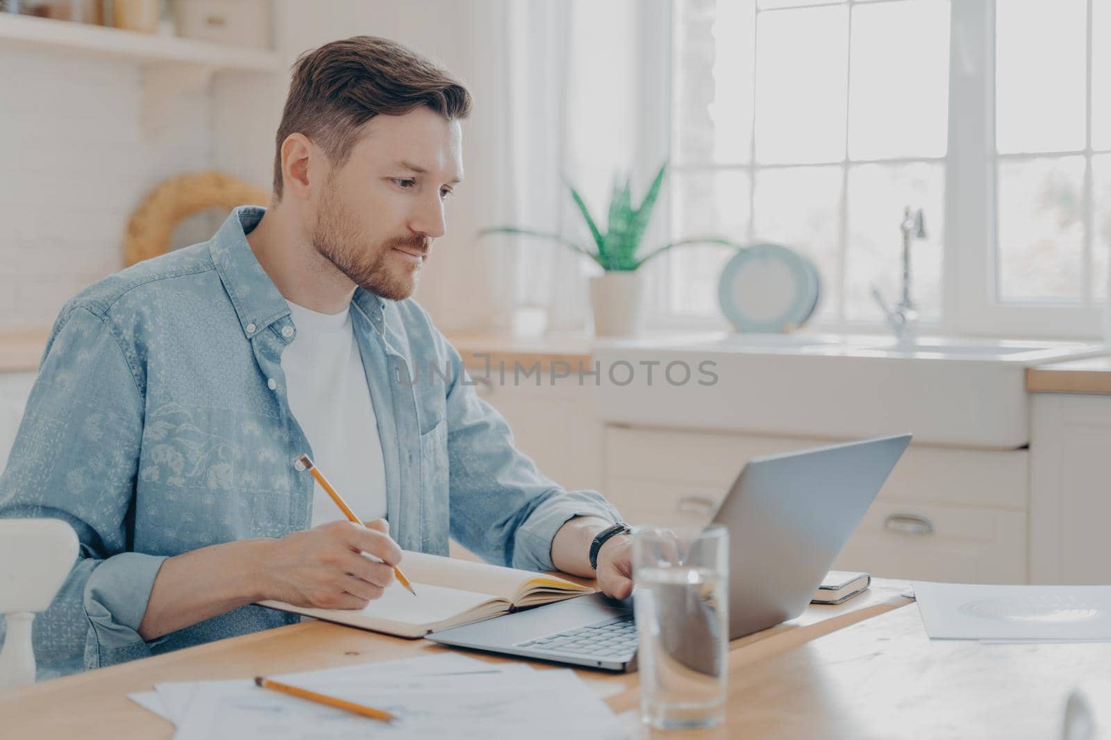 Concentrated young businessman writing notes on agenda and working remotely on laptop by vkstock