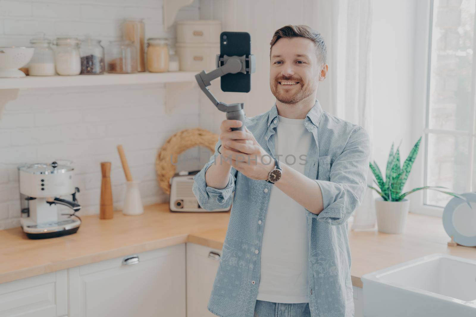 Young man blogger recording video blog or live streaming with smartphone and gimbal indoors standing in kitchen background. Social media vlogger shooting vlog, streaming online podcast on mobile phone