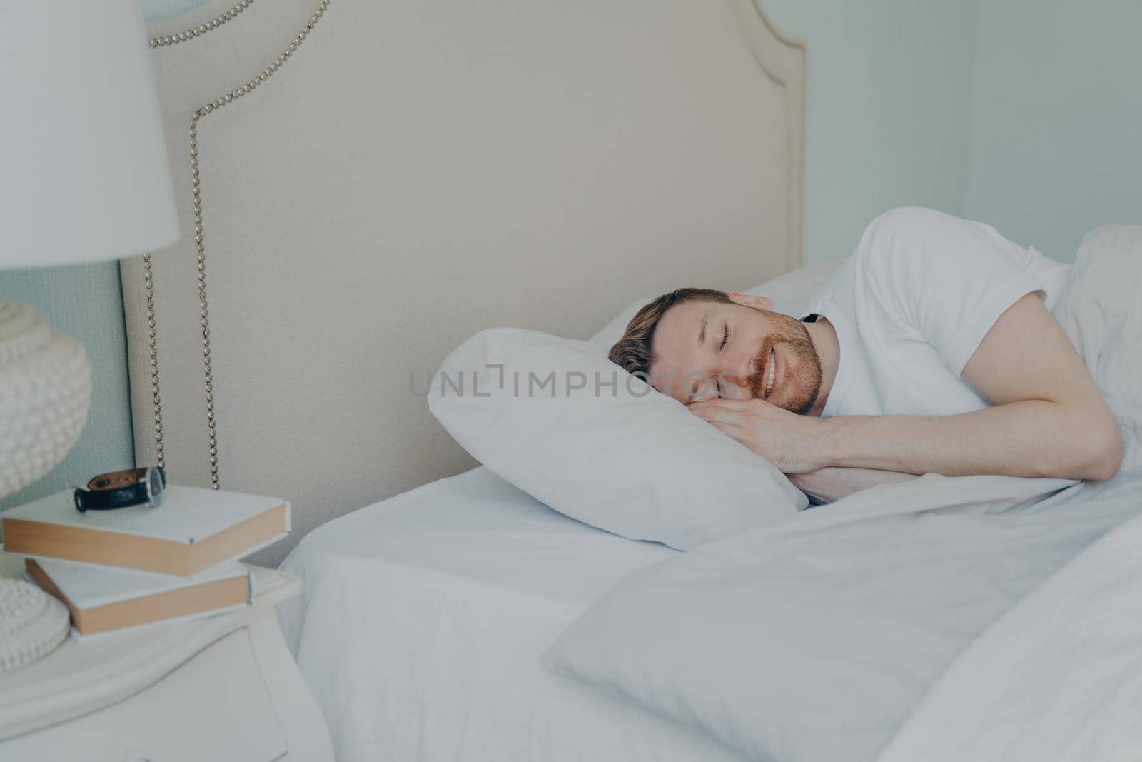 Young relaxed bearded man in casual pajama lying in bed with closed eyes, smiling and feeling happy to have rest and sleep in afternoon on day off, sweet dreaming and bedtime concept
