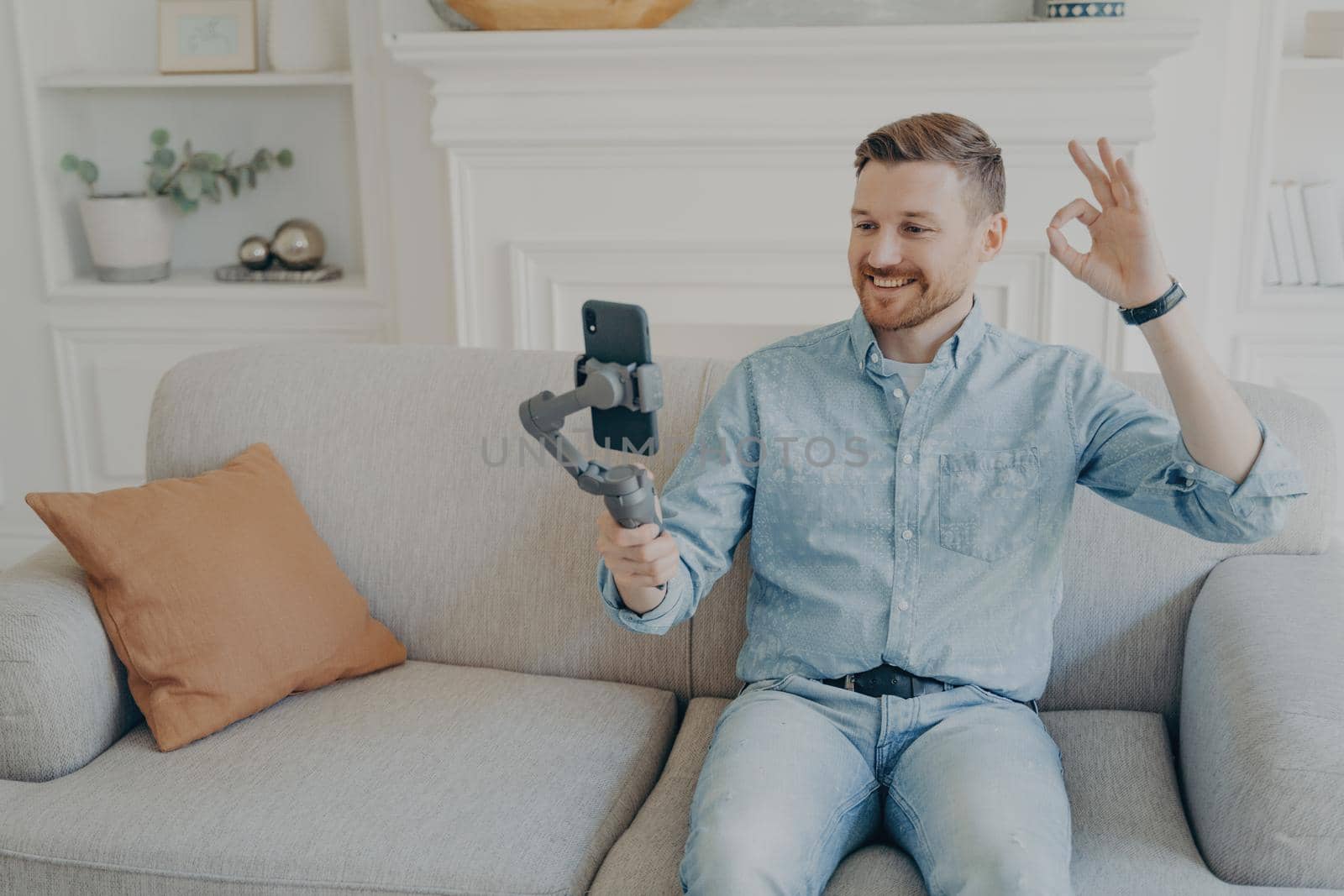 Casually dressed young man showing thumbs up while video chatting using phone attached to gimbal, sitting comfortably on beige couch in living room, having conversationg with family
