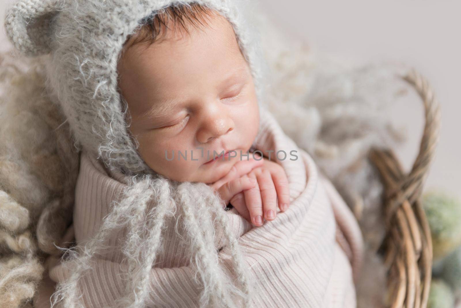 Little baby in cute white knitted beanie with ears and covered in light pink coverlet sweetly sleeping in the basket with cactus nearby