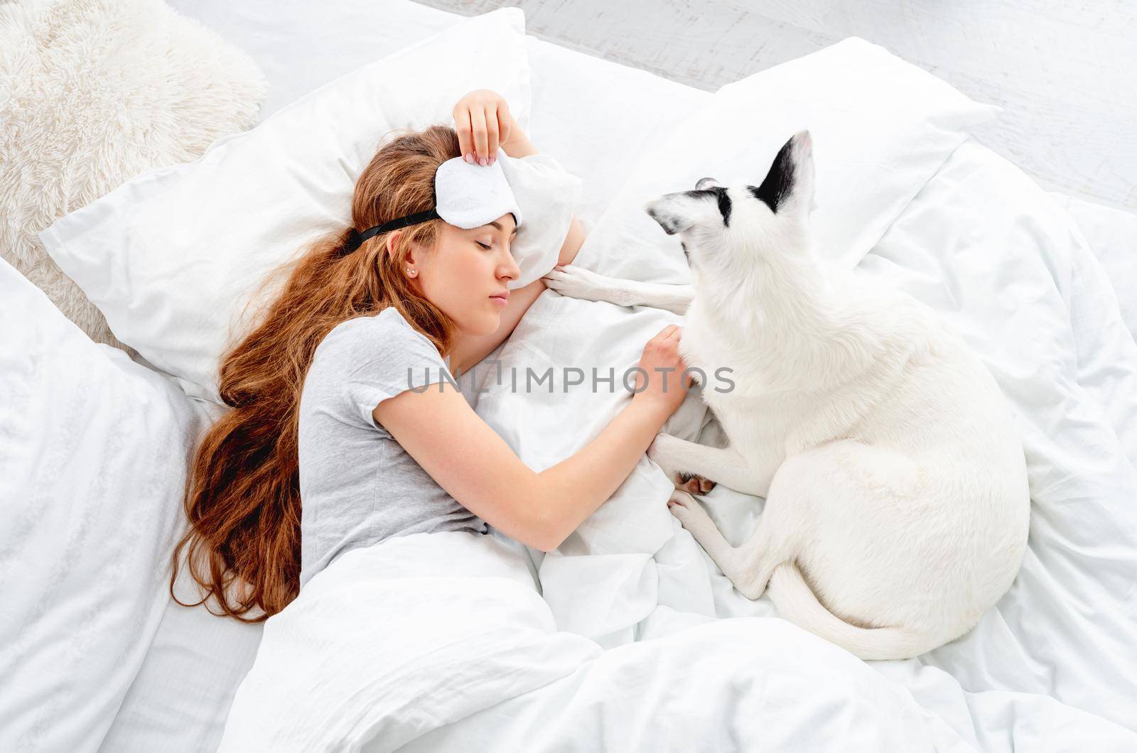 Beautiful long hair girl lying in the bed with cute dog and sleeping. Pretty young woman resting with pet. Female person wearing eye sleeping mask napping with doggy