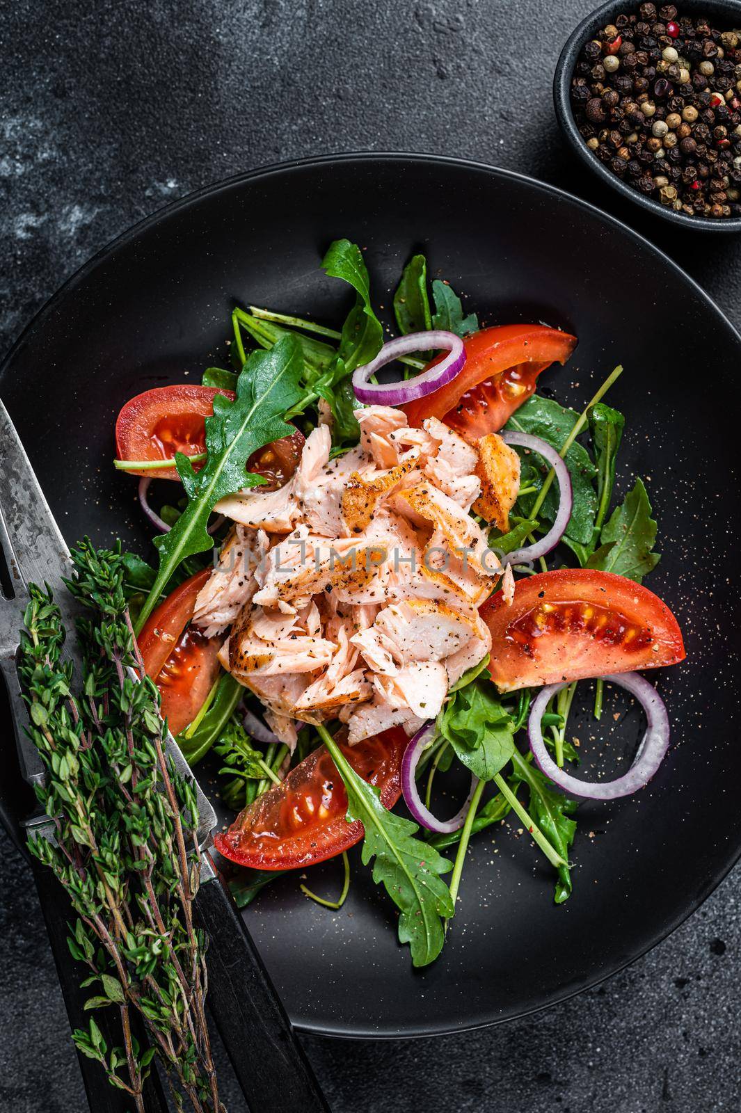 Salad with roasted salmon fillet steak, fresh salad arugula and tomato in a plate. Black background. Top view by Composter