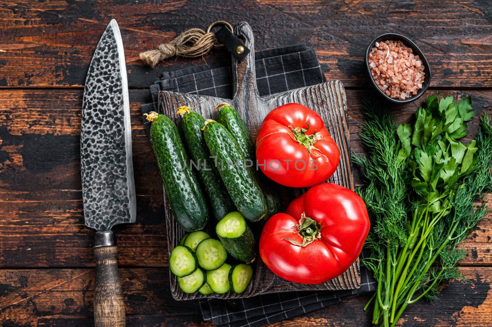 Cooking green vegetables salad with tomatoes, cucumbers, parsley, herbs. Dark wooden background. Top view.