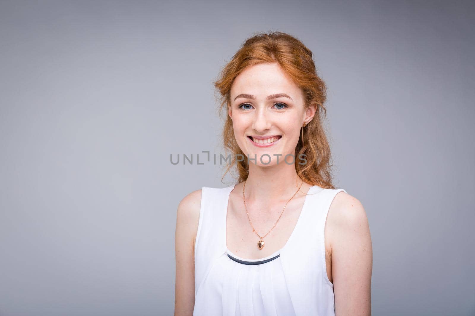 Closeup portrait young, beautiful business woman, student with lred, curly hair and freckles on face on gray background in the studio. Dressed in white blouse with short sleeves about open shoulders.