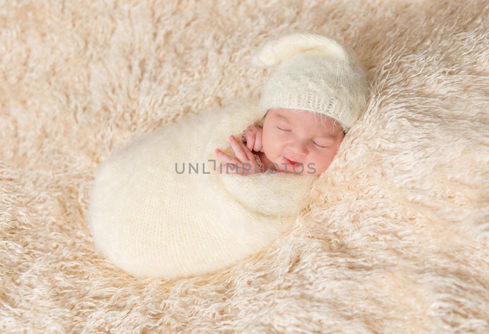 Newborn in a hat sleeping oh his side, enveloped in a soft white blanket