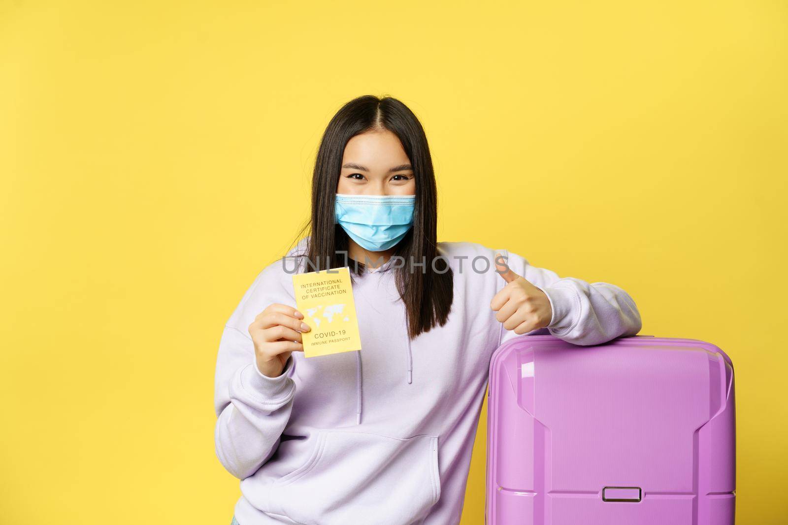 Smiling asian girl tourist in face mask, standing with suitcase, showing covid international vaccination certificate for travellers and thumbs up, yellow background.