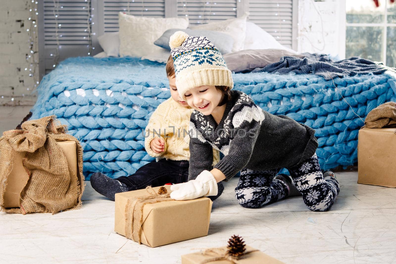 Children brother and sister sitting on floor in bedroom near bed the girl is stretching to the box with a gift on the background of Christmas decor on a sunny day.Dressed in warm knitted garment cap.