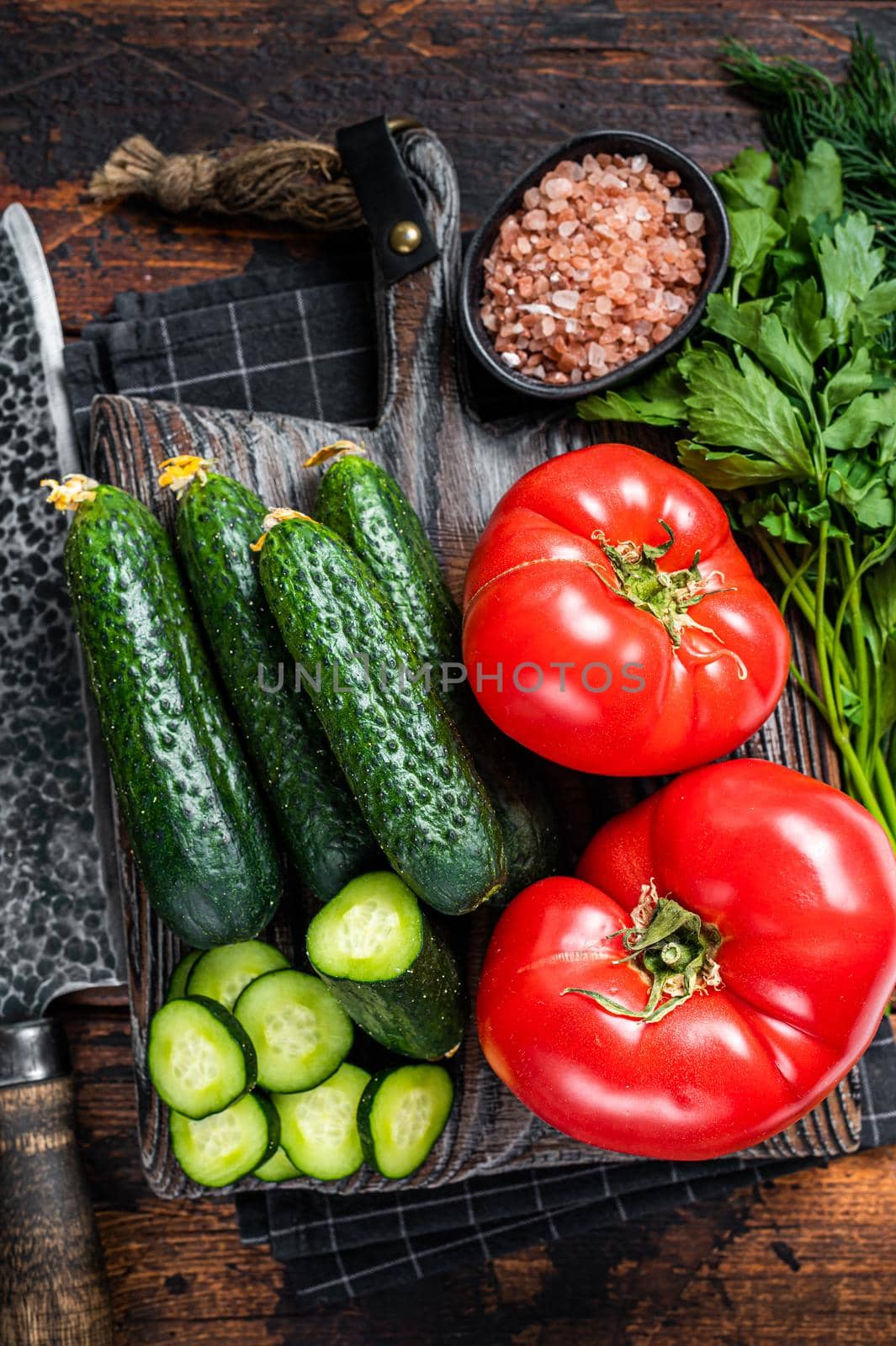 Cooking green vegetables salad with tomatoes, cucumbers, parsley, herbs. Dark wooden background. Top view.