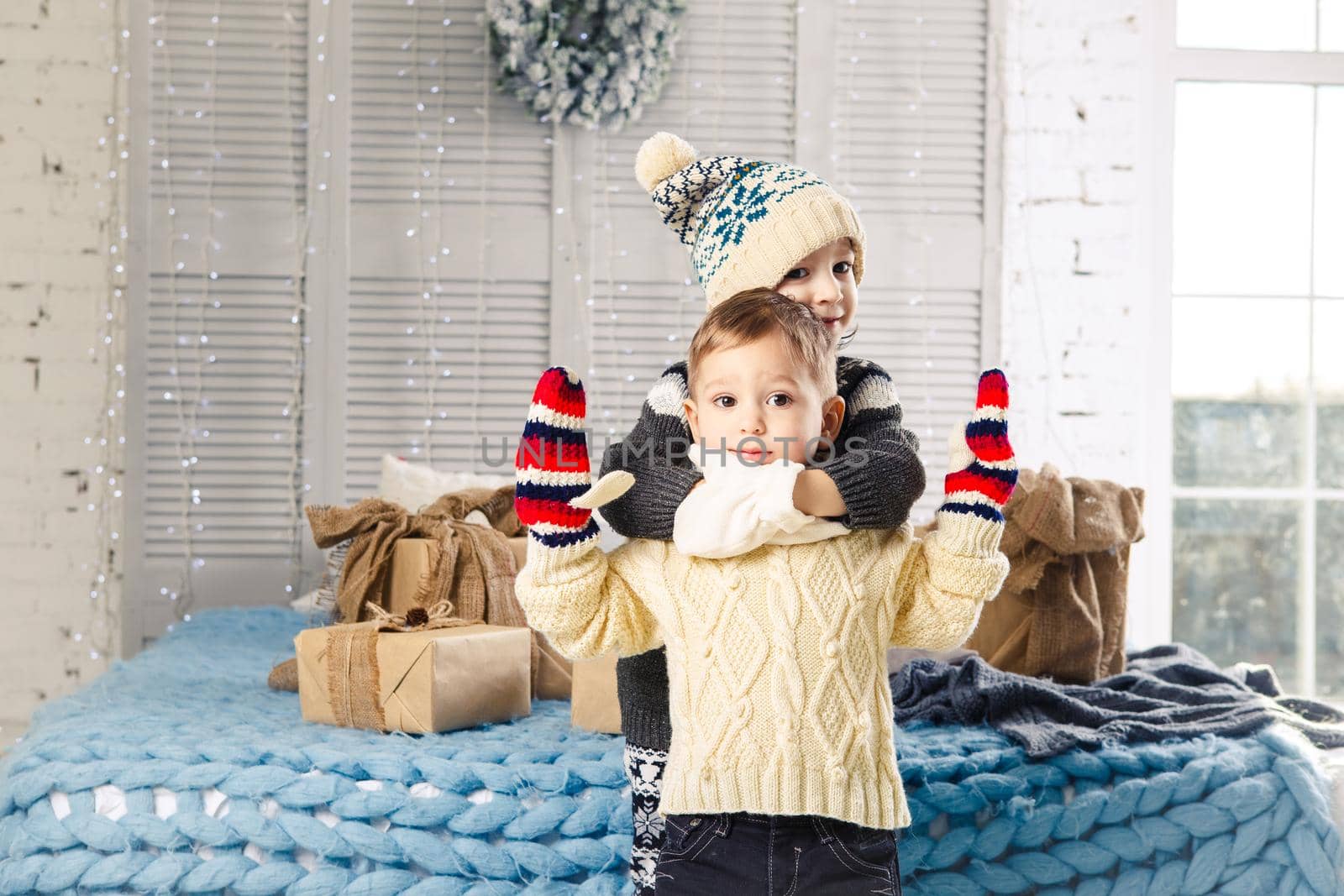 Christmas concept.Children brother and sister in embrace of the house in the bedroom near bed with boxes,gifts on back of the decor and windows on sunny day.Wear a warm woolen garment cap and mittens by Tomashevska